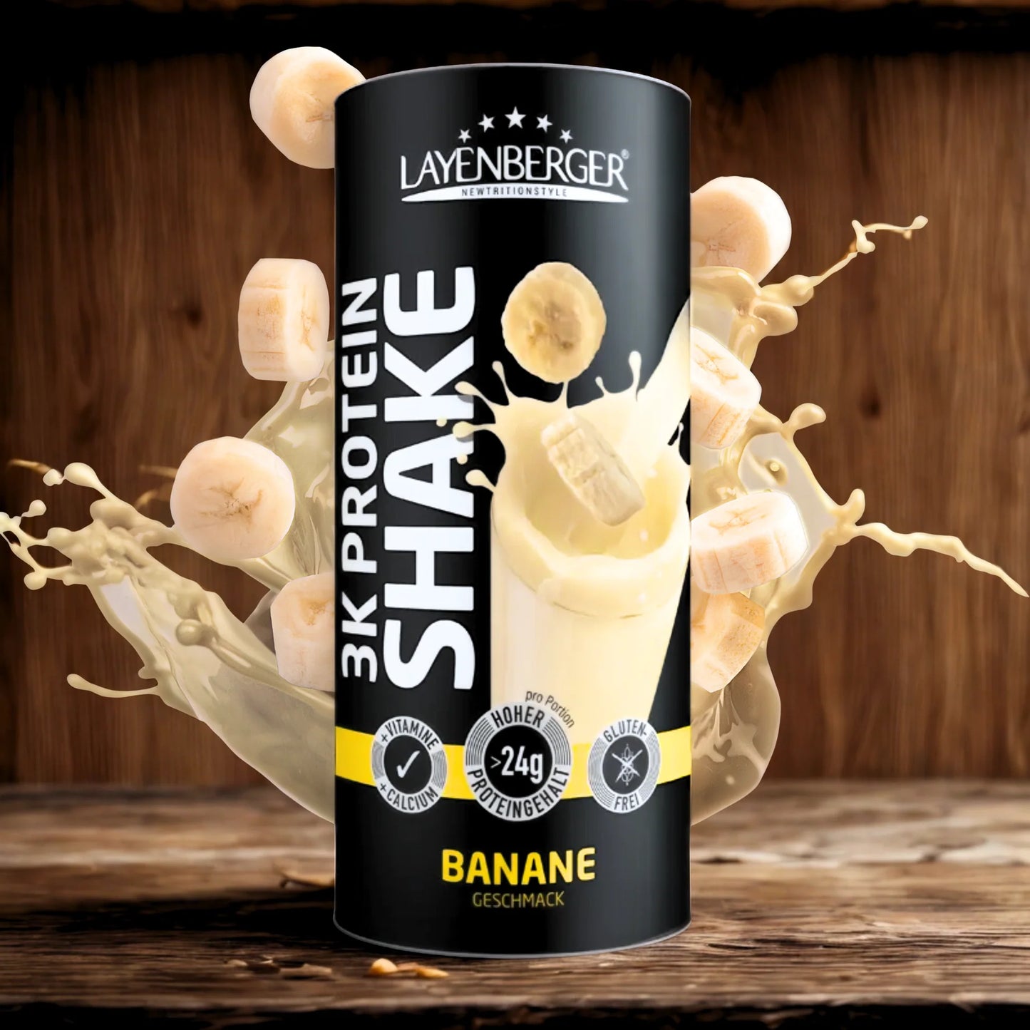 3K Protein Shake Banana; sylinterin muotoinen Layenberger 3K Protein Shake -purkki banaanin makuisena vaalealla puupinnalla; pakkaus keltainen ja valkoinen kiiltopinnalla, suljettu muovikannella; etiketissä maininnat ”glutenfrei” ja ”24 g proteiinia”; purkin ympärillä maitoroiskeita ja useita banaaniviipaleita; ei ihmisiä tai käsiä kuvassa.