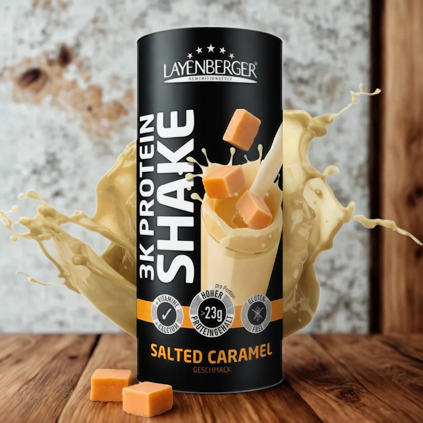 3K Protein Shake Salted Caramel; Layenberger 3K Protein Shake -tölkki suolakaramellin makuisena pystyssä; tölkki sylinterin muotoinen metallisella kiiltopinnalla; etiketissä moderni lihavoitu teksti ”Layenberger 3K Protein Shake” ja ”Salted Caramel”; ympärillä vaaleanbeigejä shakeroiskeita ja neliönmuotoisia karamellikuutioita; taustalla rustiikkia puulautoja; ei ihmisiä tai käsiä kuvassa.