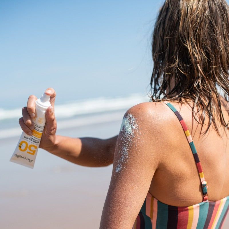 Acorelle Aurinkosuojasuihke Kasvoille ja Vartalolle SPF50: raidallisessa uimapuvussa oleva henkilö levittää SPF 50 -aurinkovoidetta olkapäälleen aurinkoisella rannalla, taustalla meren aallot; pakkaus on sylinterimäinen läppäkorkillinen, kiiltävää muovia, ja etiketissä lihavoitu "SPF 50" -teksti; kuva on kirkkaasti valaistu luonnonvalossa.