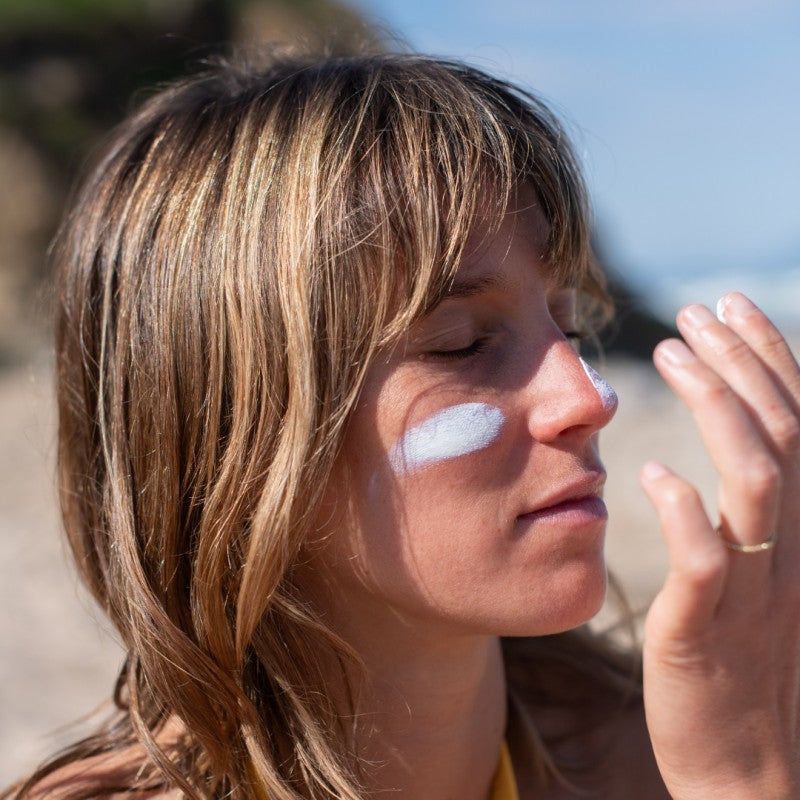 Acorelle Aurinkovoide Kasvoille SPF50: silmät kiinni oleva nainen seisoo aurinkoisella rannalla kasvoillaan valkoista aurinkovoidetta; hän on ulkona suorassa auringonvalossa, taustalla hiekkaa ja taivasta; pakkausta tai tekstiä ei näy; kuva on lähikuva muotokuvana.