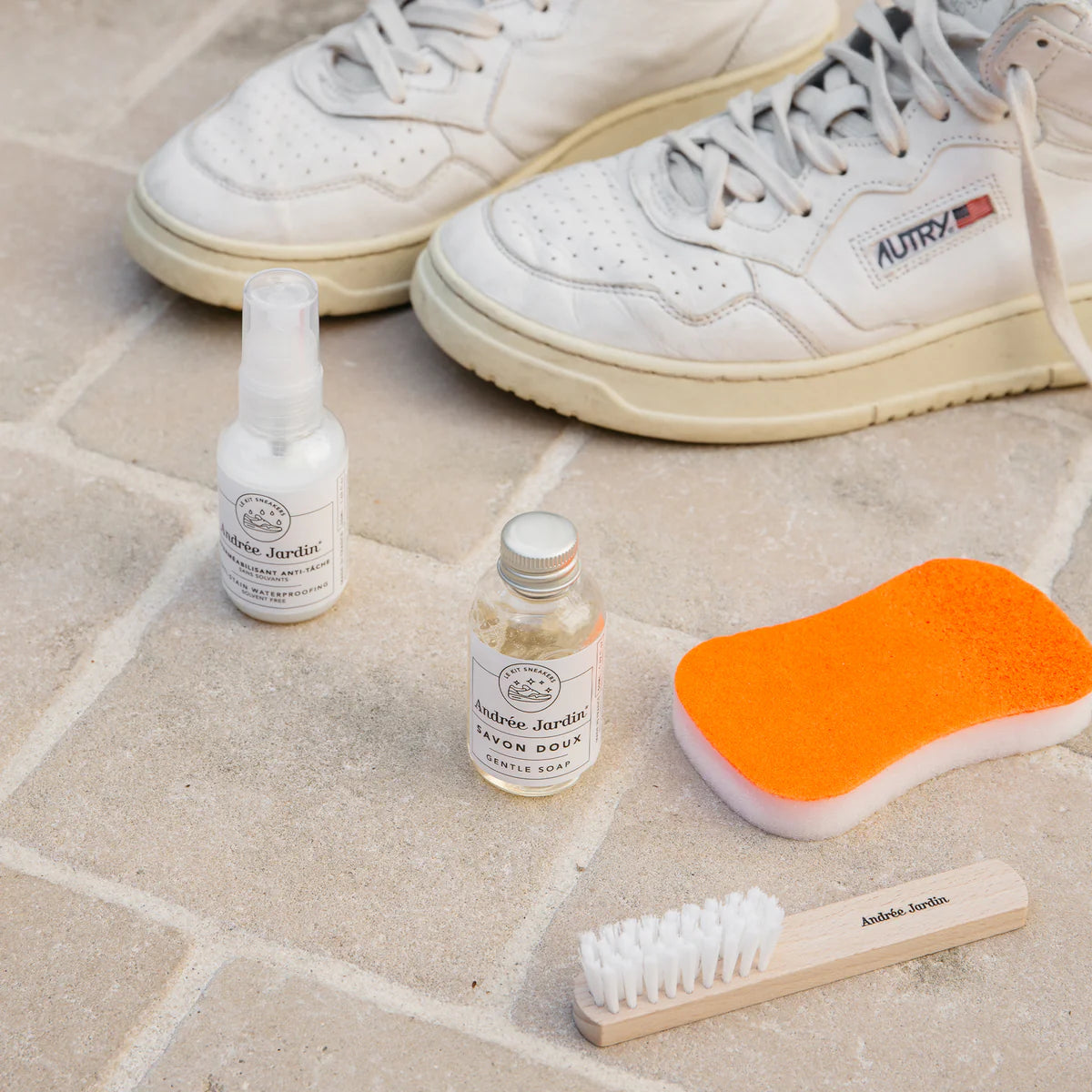 Andree Jardin Hellävarainen Kenkäsaippua; valkoiset lenkkarit vaalealla kivilattialla, edessä kenkäharja luonnonharjaksin ja puisella kädensijalla, suorakulmainen oranssi sieni, kirkas muovinen suihkupullo sekä pieni valkoinen Andrée Jardin -puhdistusainepullo; kuva otettu ylhäältä, kaikki esineet täysin näkyvissä mattapintaista taustaa vasten.