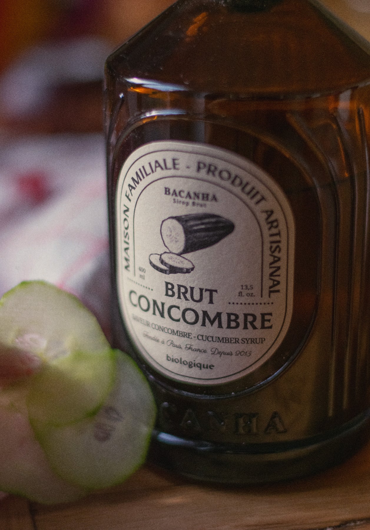 Bacanha Luomu Kurkkusiirappi; lähikuva ruskeasta pullosta, jossa lukee Brut Concombre -kurkkusiirappi, tuoreita kurkkuviipaleita vieressä puupinnalla; pullo on sylinterinmuotoinen läpikuultavasta ruskeasta lasista kiiltäväpinnalla; etiketissä tuotenimi lihavalla, groteskilla valkoisella tekstillä; viipaleet lepäävät tasaisesti pullon juuressa; ei ihmisiä tai käsiä näkyvissä; tausta on puinen pinta, kuva tiiviisti rajattu.