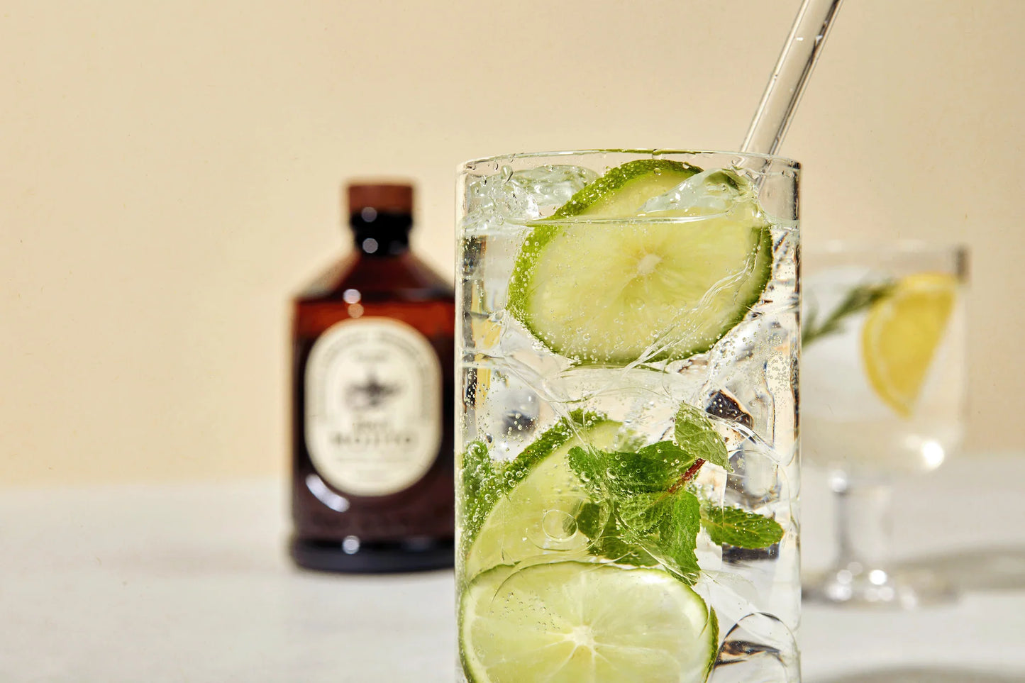 Bacanha Luomu Mojitosiirappi; lasi poreilevaa vettä limen, kurkun, mintun ja jään kera on tarjoiltu lasipillin kanssa; kirkas sylinterinmuotoinen lasi seisoo vaalealla pinnalla; taustalla pitkä ruskea pullo ilman näkyvää etikettiä sekä toinen kirkas juoma sitruunalohkon ja mintunoksan kera; ei käsiä tai ihmisiä kuvassa.