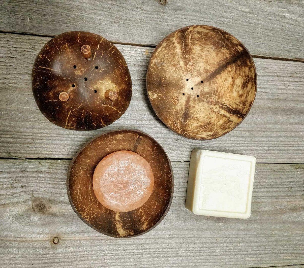 Two round coconut shell soap dishes, one with drainage holes and the other holding a pink soap, rest alongside a white bar of soap on a wooden surface. Surfaces of the dishes are matte with visible natural texture and color variations typical of coconut shells. No packaging or labels are present. Background is an unvarnished wood tabletop; no people or hands are visible in the frame.