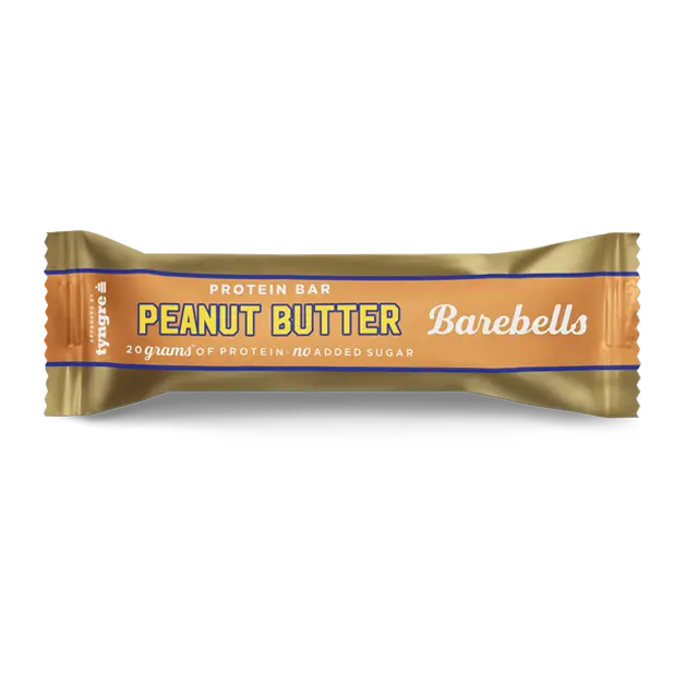Barebells Peanut Butter Proteiinipatukka; kultaväriseen kääreeseen käärittyä Barebells-proteiinipatukkaa maapähkinävoin maussa; kääreessä etusivulla korostetaan 20 grammaa proteiinia ja ei lisättyä sokeria; suorakaiteen muotoinen foliomainen pakkaus matta- ja kiiltäväpintaisin osin; brändinimi painettu lihavoiduin versaalikirjaimin; maku ilmoitettu suurella tekstillä, lisäravintotietoa alla; tausta tasainen, ei ihmisiä tai käsiä näkyvissä.