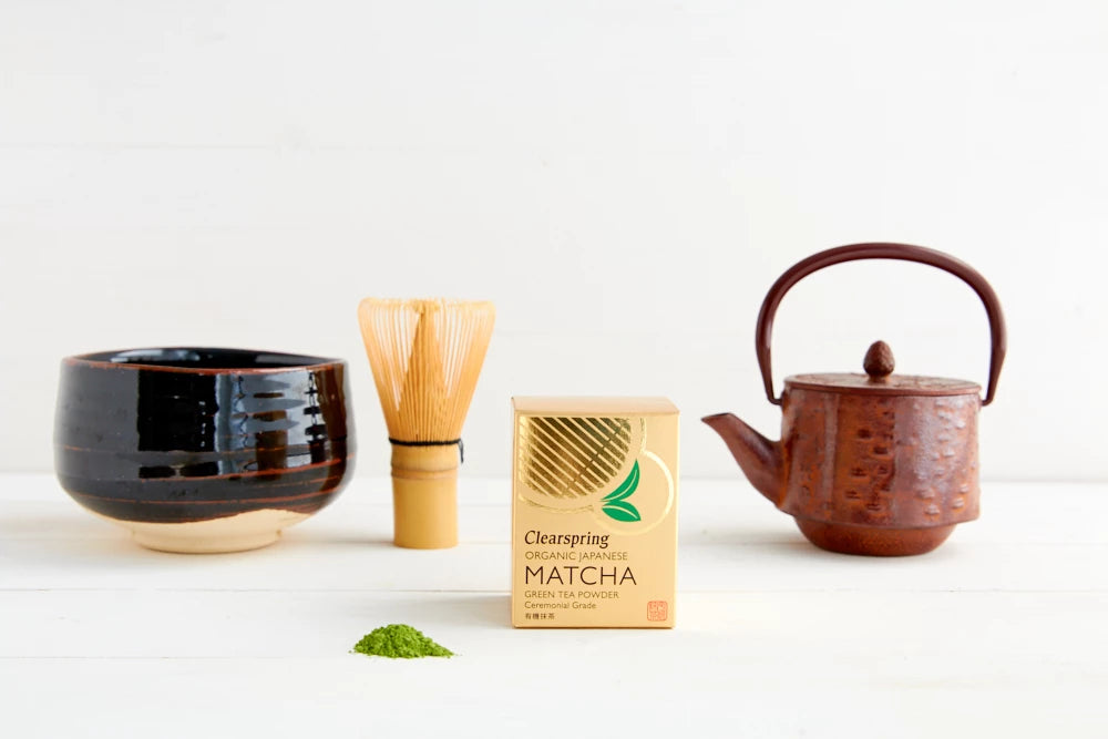 A box of Clearspring matcha powder sits between a matcha bowl, bamboo whisk, and a rustic teapot on a white surface, with a small pile of ceremonial green tea powder in front. The box is rectangular, upright, predominantly white with green accents, and matte-finished; legible label text reads “Clearspring Matcha Green Tea.” The ceramic bowl is round and light-colored. The bamboo whisk has fine prongs. The teapot has an unglazed or matte dark finish and a handle on top. No people or hands are visible; the