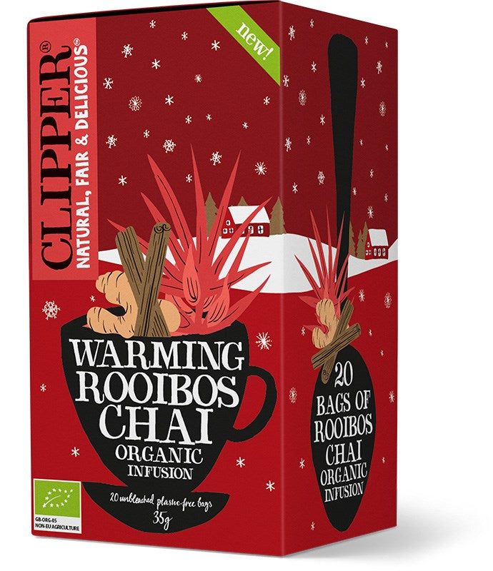 A red box of Clipper Warming Rooibos Chai Organic Infusion tea, featuring a black teacup with cinnamon sticks, star anise, and red leaves. The packaging is rectangular with a matte finish. White sans-serif text identifies the organic product. Background includes illustrations of stylized snow and houses; no people or hands are visible in the image.