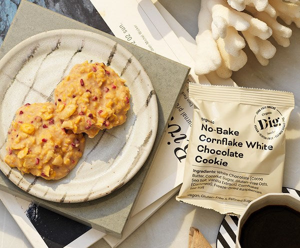 Dig Luomu Cornflake-Valkosuklaakeksi; kaksi paistamatonta cornflake-valkosuklaakeksiä lepää mattapintaisella keramiikkalautasella suorakulmaisen tuotepakkauksen vieressä, jossa lohko- ja kaunokirjoitusfontteja, aseteltuna pinottujen kovakantisten kirjojen kansille kuvioidulla koristepöydällä; kuva otettu ylhäältä ja sisältää vain esineitä, ei ihmisiä tai käsiä näkyvissä.