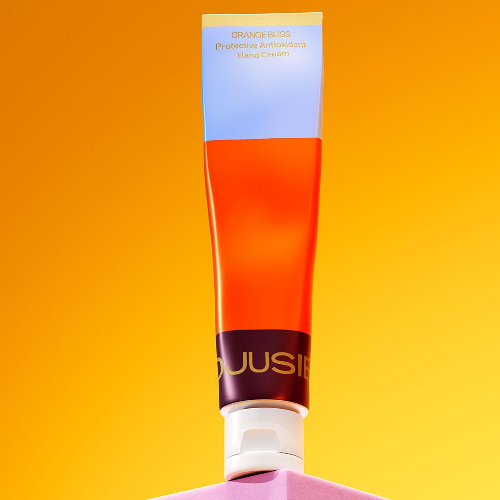 A tube of Orange Bliss Protective Antioxidant Hand Cream stands upright on a pink surface, set against a vibrant citrus-inspired yellow-orange background. The tube is cylindrical with an opaque, smooth finish and a screw cap. The label displays the product name in clear, sans-serif font. No people or additional objects are present in the shot.