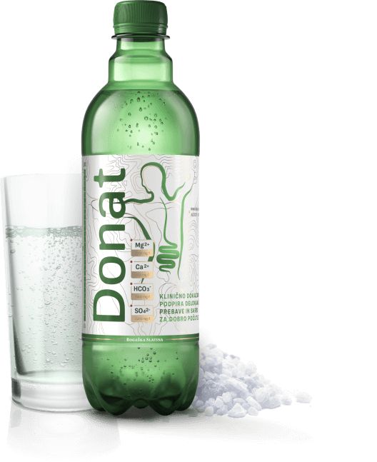 A green glass bottle labeled “Donat” is shown upright on a flat surface, next to a cylindrical clear glass containing sparkling mineral water with visible bubbles. Small white mineral crystals are scattered nearby. The bottle has a glossy finish and the label features clear, bold sans-serif text. The scene is evenly lit with no people or hands present. A green bottle labeled Donat stands next to a glass filled with sparkling mineral water and some white mineral crystals on the surface.