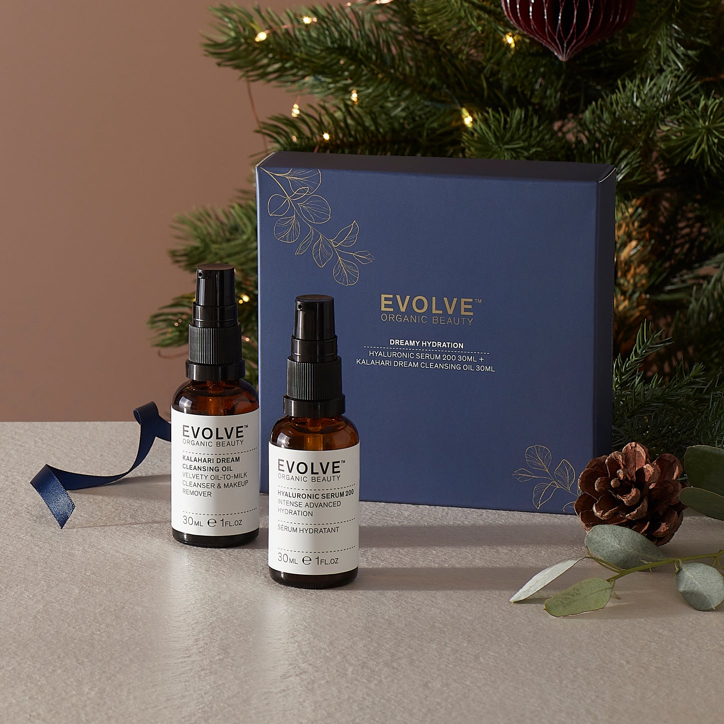 Evolve Dreamy Hydration; kaksi Evolve Organic Beauty -seerumipulloa valkoisin etikettein ja mustin pumppukansin seisoo pystyssä; etiketissä luettavissa brändinimi modernilla groteskifontilla; pullot sinisen mattapintaisen suorakaiteen muotoisen lahjarasian vieressä; beigen sävyinen tasainen alusta alla; sivussa näkyy osa joulukuusesta pyörein koristein ja käpyin; ei ihmisiä tai toimintaa, studiotyylinen asetelmakuva.