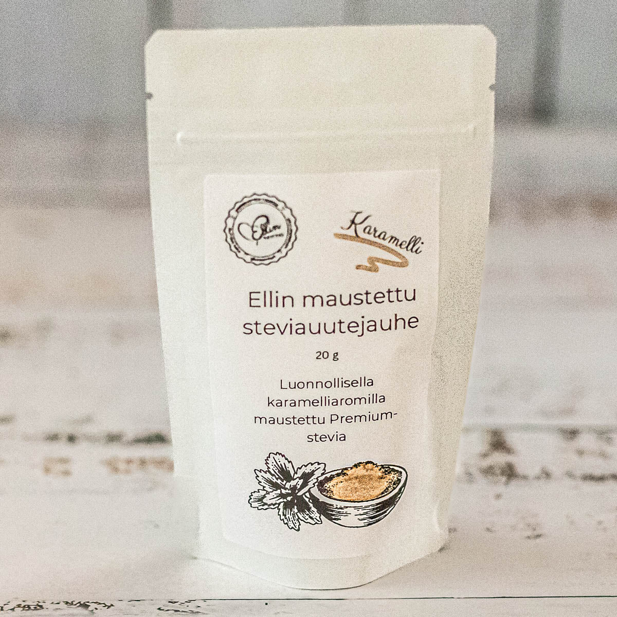 Ellin Karamellistevia; valkoinen, mattapintainen joustava pussi, jossa lukee "Ellin maustettu steviauuttejauhe" ja mainitaan karamellin maku, seisoo pystyssä vaaleaa puukuvioitua pintaa vasten; pakkauksen kuvituksena vihreä stevialehti ja karamellipaloja; typografia on modernia groteskia, painettu mustalla ja oranssilla; ei ihmisiä tai käsiä näkyvissä.