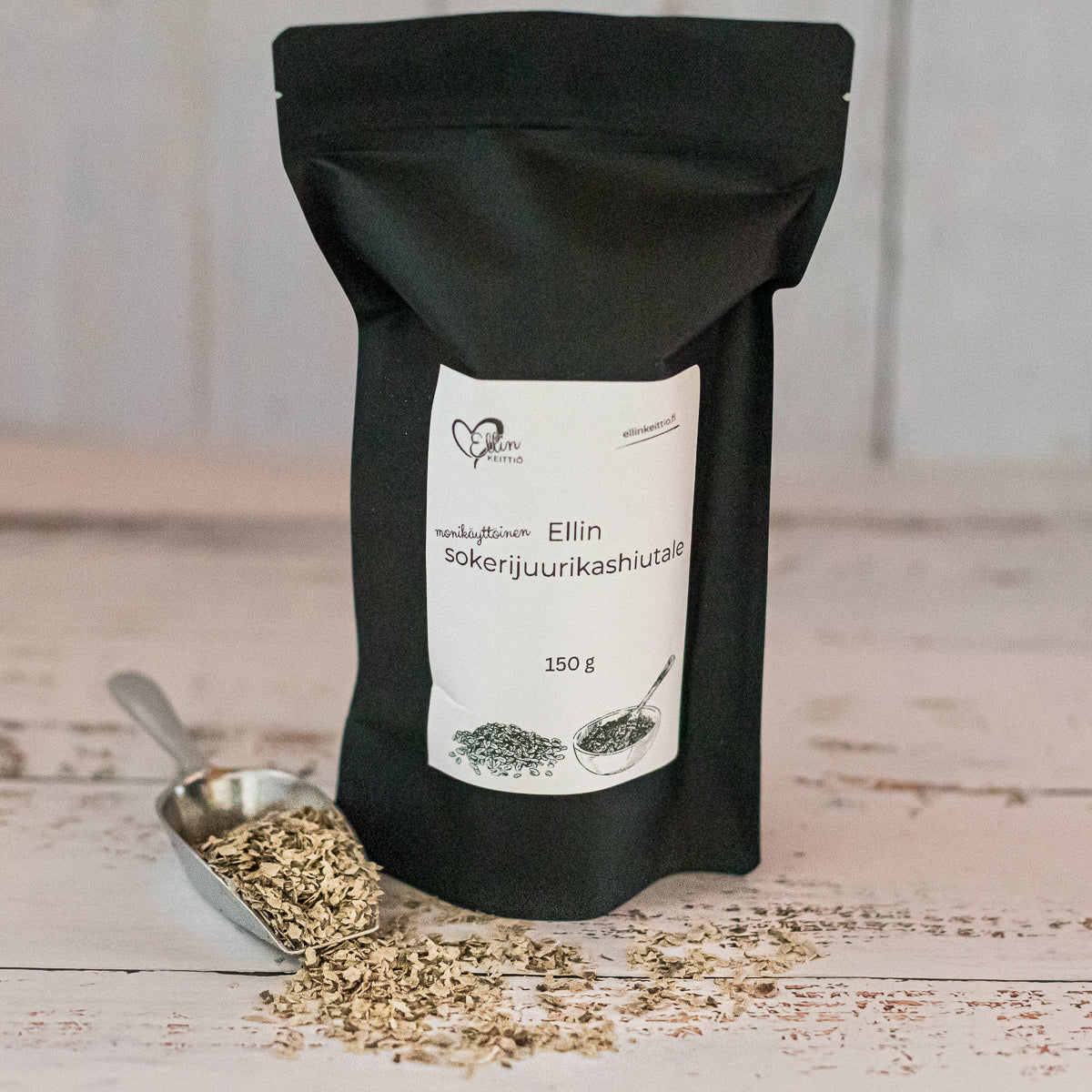 Ellin Sokerijuurikashiutale; musta uudelleensuljettava pussi, jossa lukee Ellin sokerijuurikashiutale 150g, seisoo pystyssä hopeisen metallilusikan vieressä, jossa vaaleita, epäsäännöllisiä hiutaleita; pussi on mattapintainen valkoisella painetulla tekstillä; molemmat esineet vaakasuuntaisilla valkoisilla puulaudoilla, kuva otettu hieman ylhäältä; ei ihmisiä tai käsiä näkyvissä.