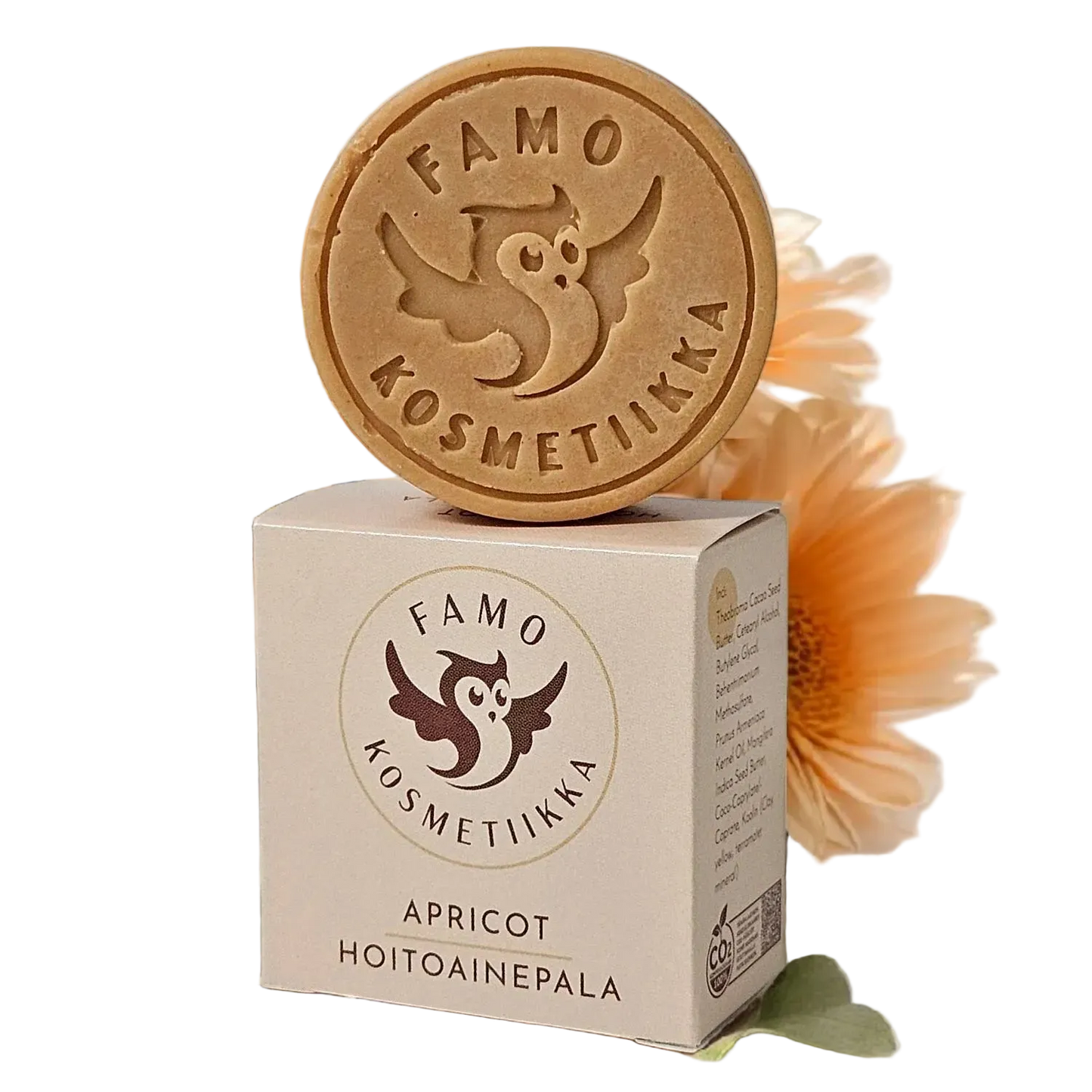Famo Apricot Hoitoainepala: pyöreä Famo Kosmetiikka -aprikoosisaippua rasiansa päällä, taustalla kukka; saippua on kiinteä, vaaleanoranssi ja mattapintainen, kohokuvioitu pöllölogo; suorakulmainen rasia on valkoinen suomenkielisellä tekstillä ja samalla pöllölogolla, lihavoitu groteskikirjasin; ei ihmisiä tai käsiä näkyvissä, kuva otettu ylhäältä.