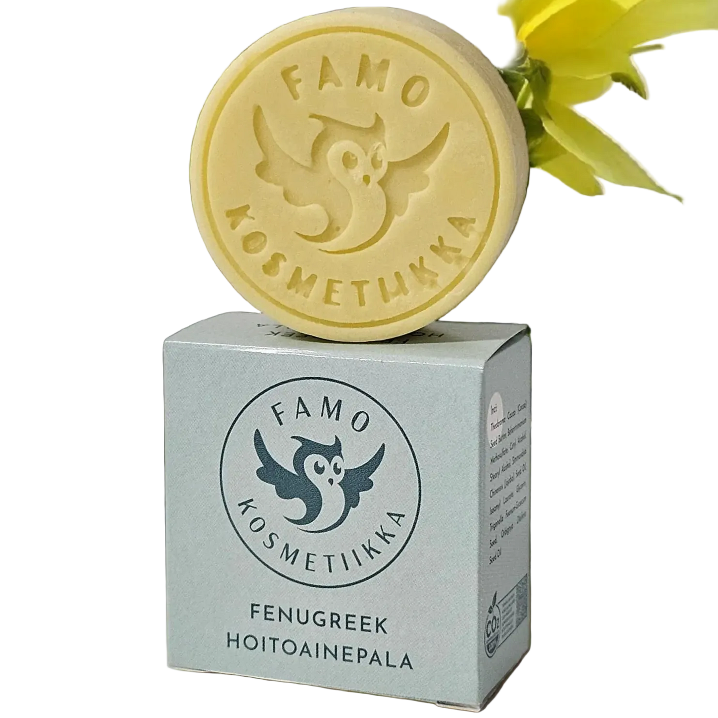 Famo Fenugreek Hoitoainepala: pyöreä keltainen saippuapalanen pöllölogolla ja Famo Kosmetiikka -tekstillä lepää sitä vastaavan suorakulmaisen rasian päällä, jossa teksti "Famo Kosmetiikka Fenugreek Hoitoainepala" lihavoidulla groteskikirjasimella; sekä palasella että rasialla mattapinta; taustalla keltaisia kukkia; ei ihmisiä tai käsiä näkyvissä.