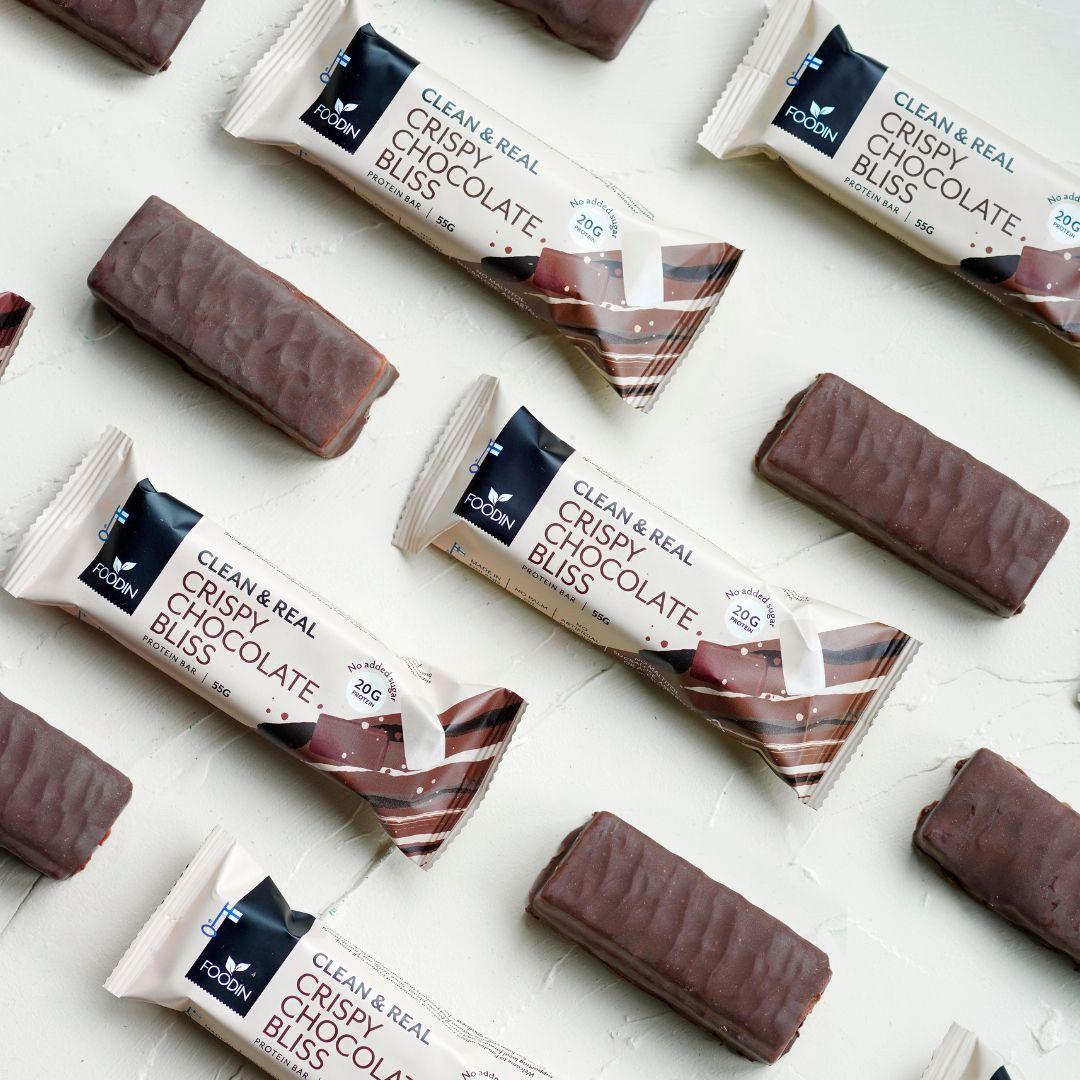 Foodin Clean & Real Protein Bar Crispy Chocolate Bliss; suklaapatukoita, sekä kääreettöminä että pakkauksessa merkittynä "Crispy Chocolate Bliss", aseteltuna siistiin, vinoittaiseen kuvioon vaalealle taustalle; suorakulmaiset patukat tekstuurisella suklaapäällysteellä puolikiiltävällä pinnalla; pakkaus suorakulmainen näkyvällä typografisella etikettitekstillä; ei ihmisiä eikä käsiä näkyvissä; kuva ylhäältä.
