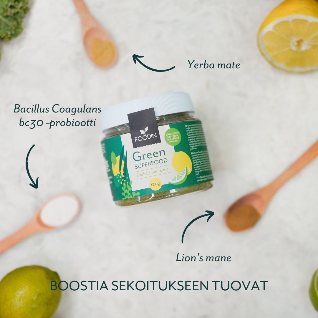 A jar of Foodin Green Superfood is surrounded by lemons, a lime, wooden spoons, and white powder. The jar is cylindrical with a matte black lid; the label features clear sans-serif text and green accents. Arrows point to labeled ingredients: yerba mate, lion’s mane, Bacillus Coagulans bc30 -probiotti. Text reads: BOOSTIA SEKOITUKSEEN TUOVAT.. The background is plain white. No people or hands are present. Shot is from above.