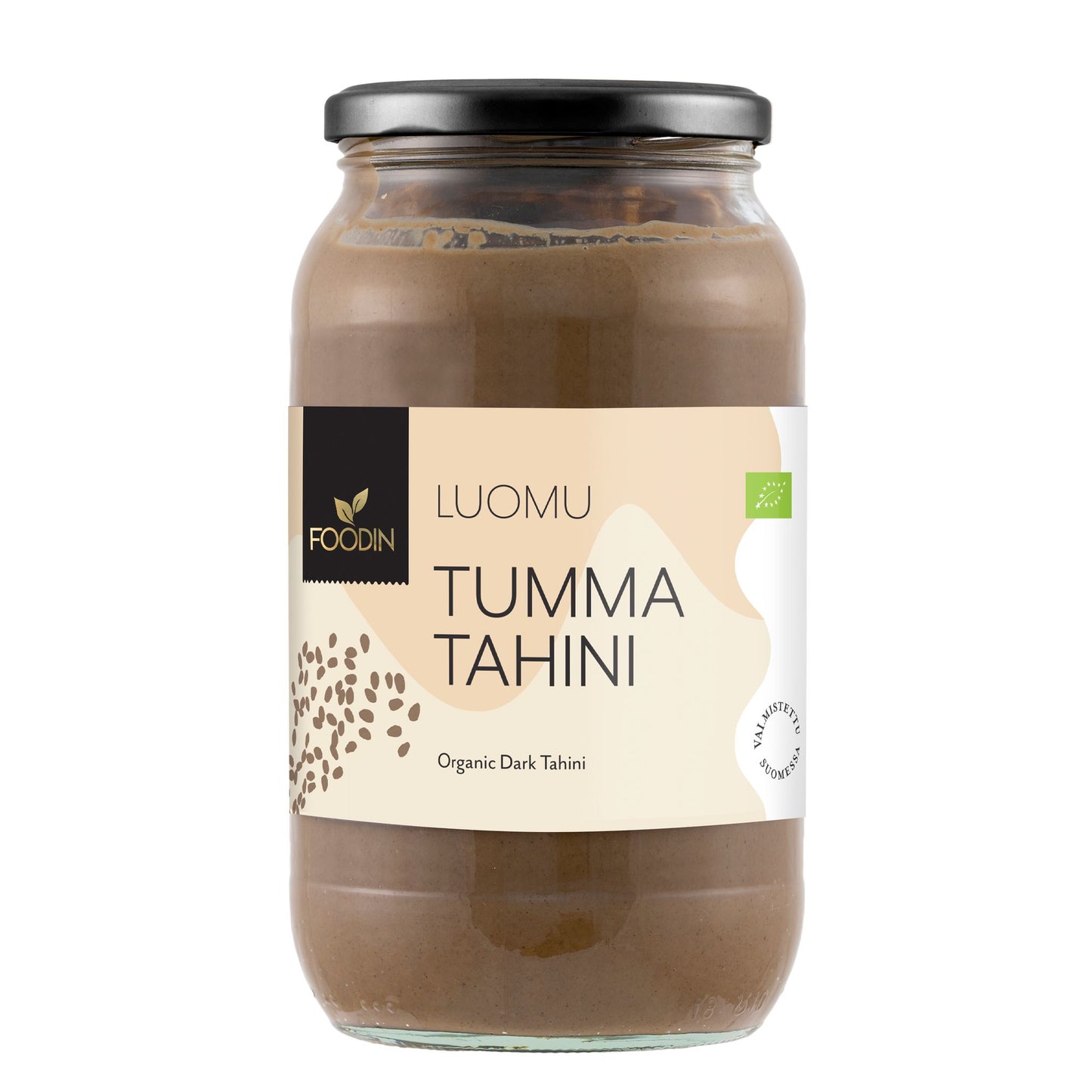 Foodin Luomu Tahini Tumma -lasipurkki; musta muovinen kansi; sylinterimäinen purkki, jota kiertää mattapintainen beige-ruskea etiketti tekstillä "Luomu Tumma Tahini" selkeällä tekstausvapaalla kirjasimella; kuvattu silmän korkeudelta yksiväristä taustaa vasten.