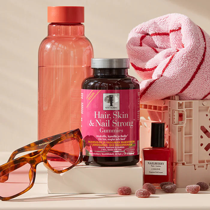 A bottle of Hair, Skin & Nail Strong Gummies with a clear plastic body and purple cap displays bold white and pink label text. The pink towel is folded with a soft fabric texture. Red nail polish comes in a small glass bottle with a shiny finish. Black sunglasses have dark lenses and plastic frames. Several peach-colored gummies are scattered nearby. There is also a tall clear water bottle. All items are arranged on a smooth flat surface against an even peach-colored background under soft lighting, shot