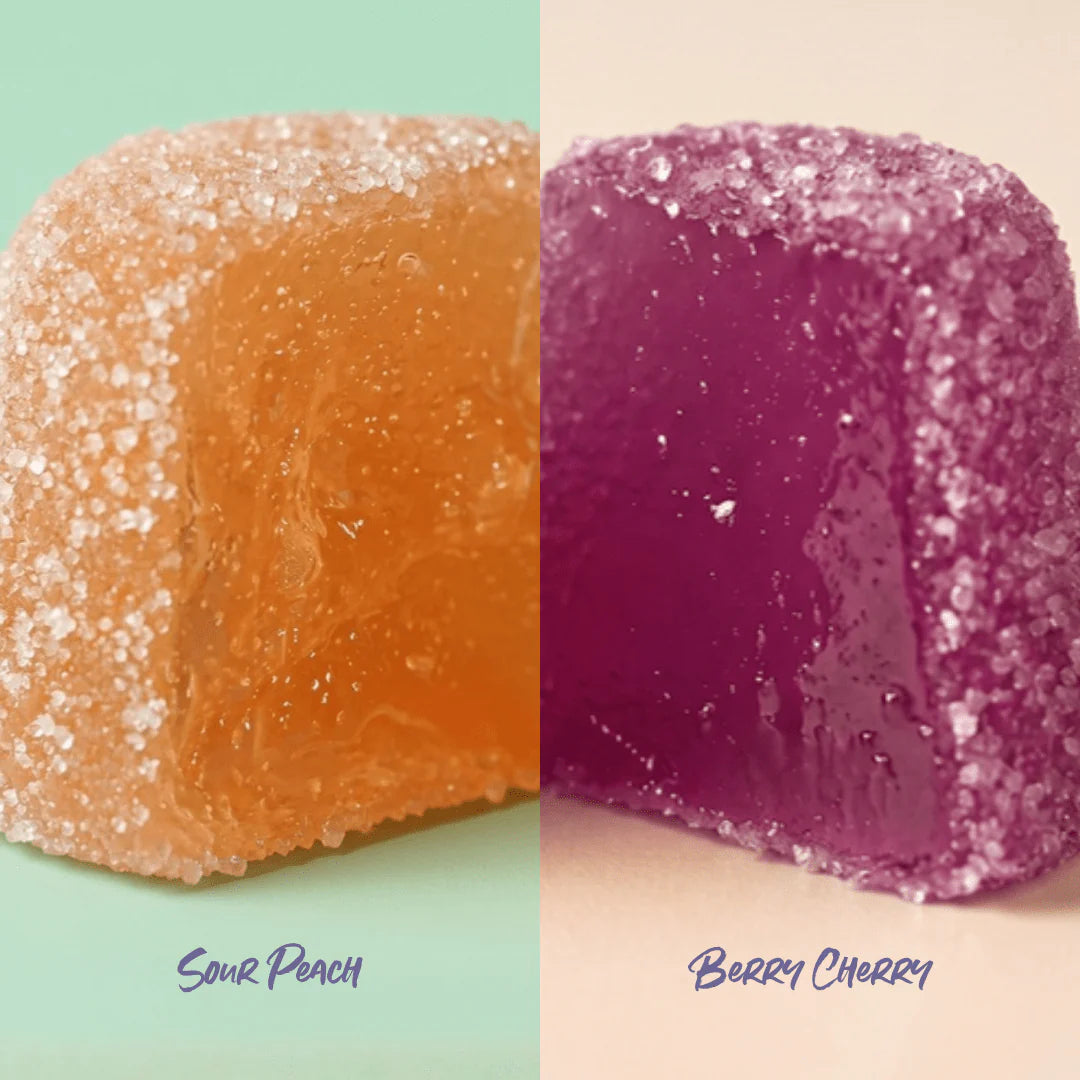 Lähikuva kahdesta sokerikuorrutetusta hyytelömakeisesta, joista oranssissa lukee Sour Peach vasemmalla ja violetissa Berry Cherry oikealla, pehmeitä pastellitaustoja vasten. Molemmat makeiset ovat pyöristettyjä neliöitä, joiden pinnalla on rakeinen, mattainen sokerikuorrutus. Etiketeissä käytetään yksinkertaista pääteviivatonta fonttia painettuna suoraan makeisen päälle tai lähelle. Pakkausta ei näy. Ihmisiä tai käsiä ei näy kuvassa.