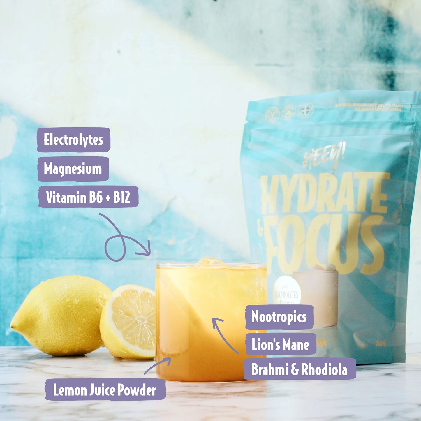 Heey! Hydrate & Focus Electrolytes, maku Lemon; lasillinen keltaista juomaa sitruunan kanssa, etiketöity ainesosin kuten elektrolyytit, vitamiinit ja nootropit, etualalla, taustalla Hydrate & Focus -juomajauhepussi; molemmissa vaaleanvioletit etiketit erityisominaisuuksin; pakkaus yksittäisannos, pussissa modernia groteskitekstiä; ei ihmisiä tai käsiä; siisti studioasetelma, kuvattu suoraan edestä.
