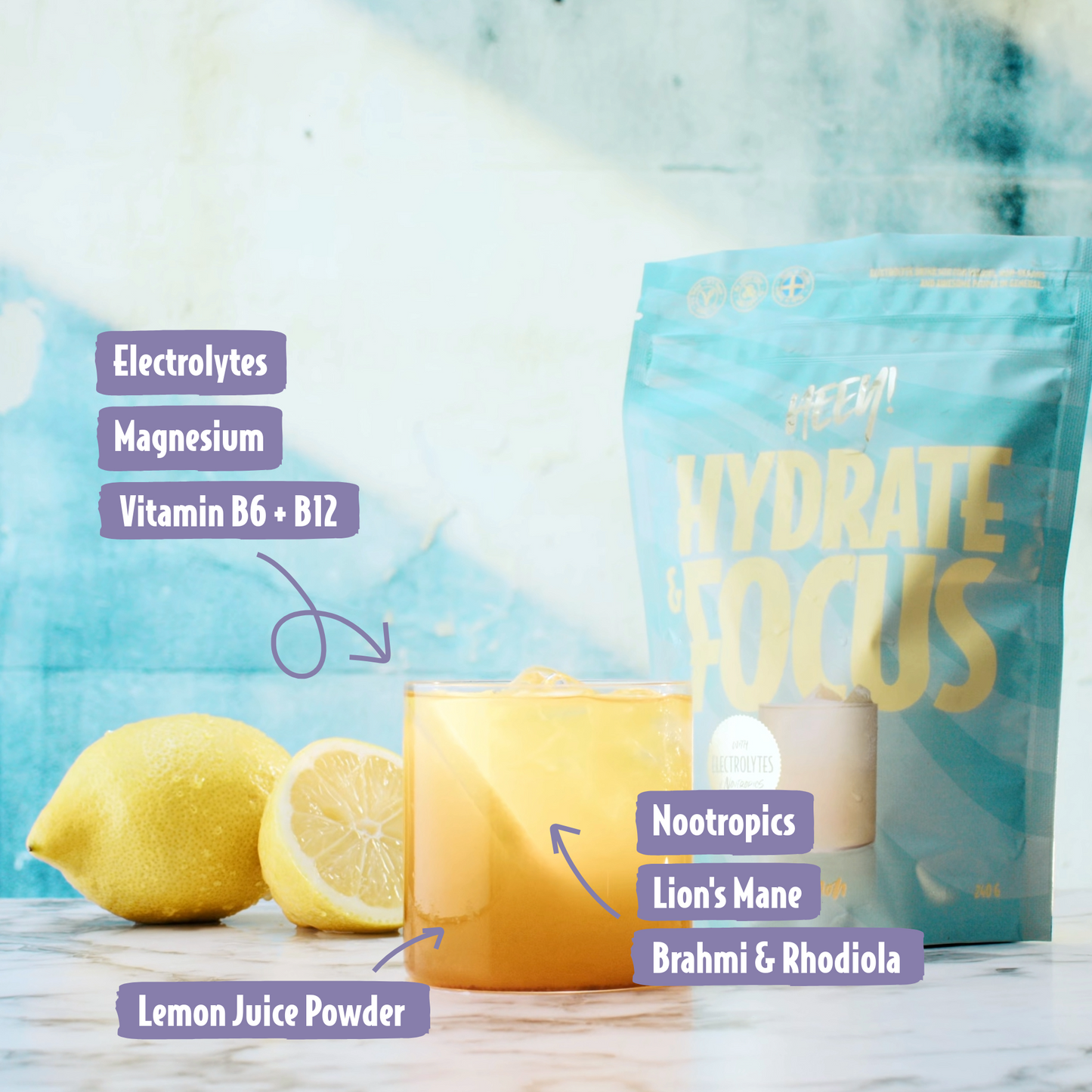 Heey! Hydrate & Focus Electrolytes, maku Lemon; lasillinen keltaista juomaa sitruunan kanssa, etiketöity ainesosin kuten elektrolyytit, vitamiinit ja nootropit, etualalla, taustalla Hydrate & Focus -juomajauhepussi; molemmissa vaaleanvioletit etiketit erityisominaisuuksin; pakkaus yksittäisannos, pussissa modernia groteskitekstiä; ei ihmisiä tai käsiä; siisti studioasetelma, kuvattu suoraan edestä.