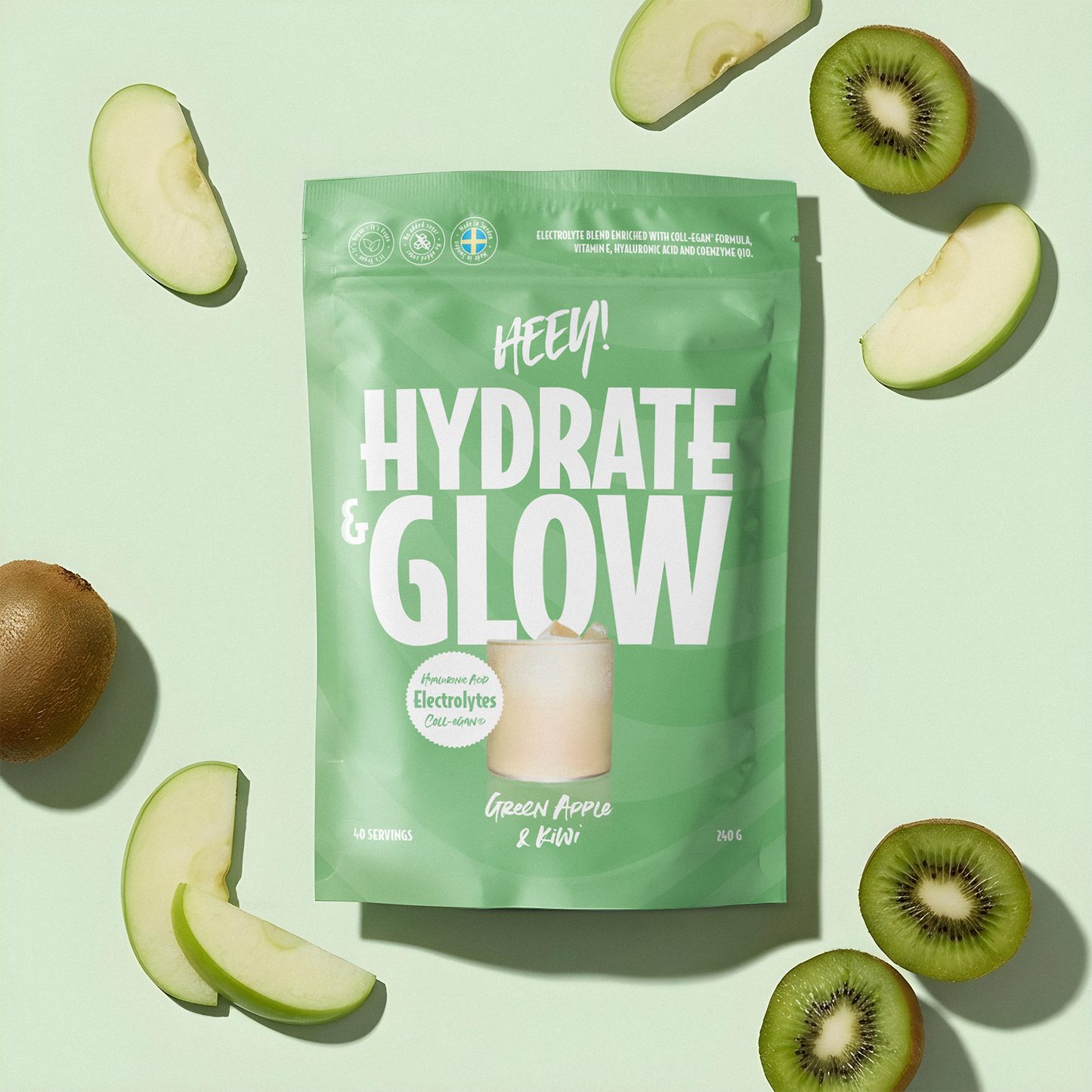 Vihreä Hydrate & Glow -juomajauhepussi on aseteltu vaalealle pinnalle, ympärillä viipaloituja vihreitä omenoita ja kiivejä. Pussi on mattapintainen, suorakulmainen ja tiivistetyllä yläreunalla. Lihavoitu valkoinen pääteviivaton typografia näyttää tekstin Hydrate & Glow ja mainitsee elektrolyytit nesteytyksen tueksi sekä maun green apple & kiwi. Ihmisiä tai käsiä ei näy kuvassa. Kuva on otettu ylhäältä.