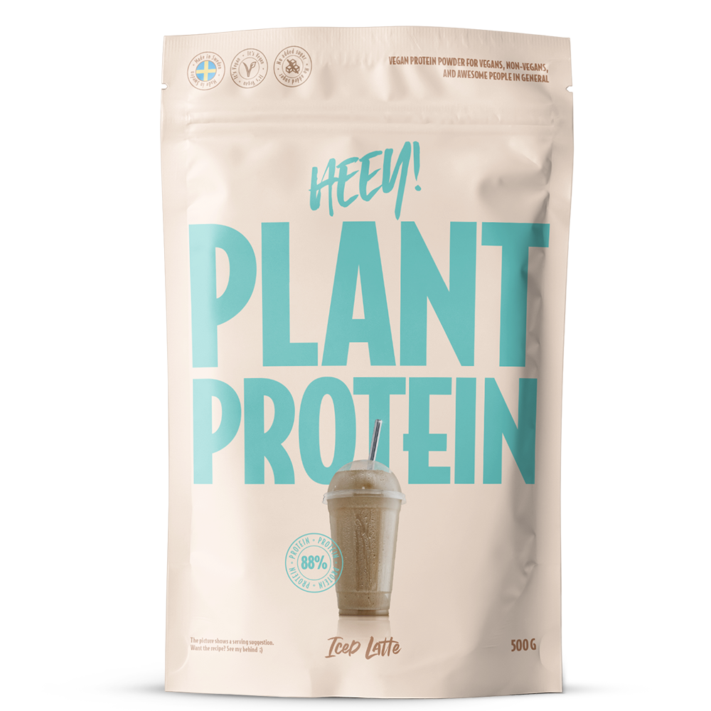 Heey! Plant Protein Iced Latte; beige 500 g:n pussi, jossa lukee HEEY! PLANT PROTEIN suurin sinivihrein kirjaimin, kuvituksena kylmä latte kupissa pillin kera etupuolella; pussin pinta on mattapintainen, muoto suorakulmainen pyöristetyin reunoin; pakkauksen teksti mainitsee vegaaniset ainesosat ja korkean proteiinipitoisuuden; ei ihmisiä tai käsiä näkyvissä, kuva on pelkkä tuotekuva neutraalia taustaa vasten.