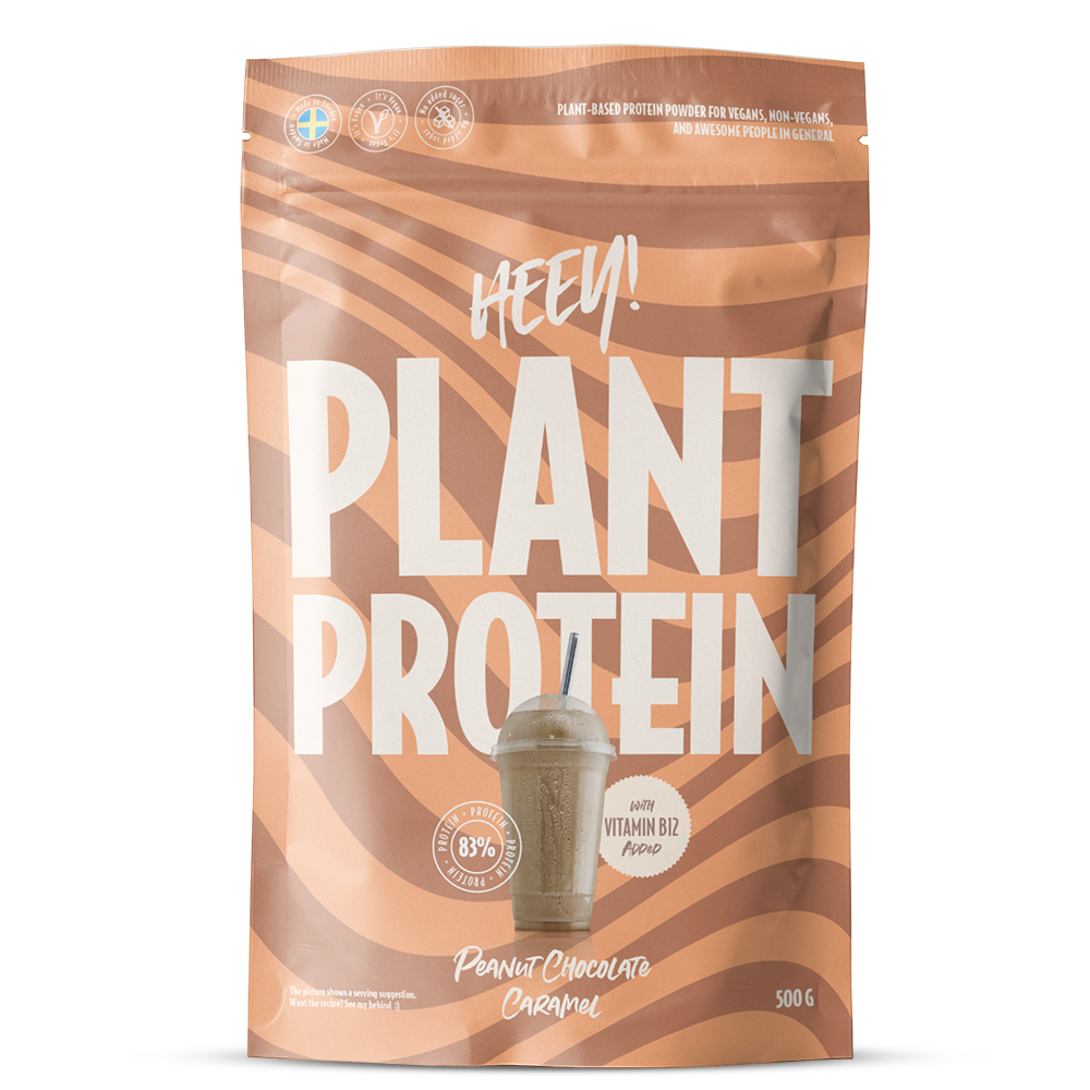 Heey! Plant Protein Peanut Chocolate Caramel; beige ja ruskea pussi, jossa lukee HEEY! PLANT PROTEIN, kuvassa smoothie, 83 % proteiinia -merkintä ja maku Peanut Chocolate Caramel; pakkaus korostaa kasvipohjaista B12-vitamiinia ja vegaanista proteiinipitoisuutta; mattapintainen pussi lohkomaisella valkoisella groteskitekstillä merkin nimessä, lisätekstit ja kuvitus valkoisena ja ruskeana; studiokuva yksivärisellä taustalla, ei ihmisiä tai rekvisiittaa näkyvissä.
