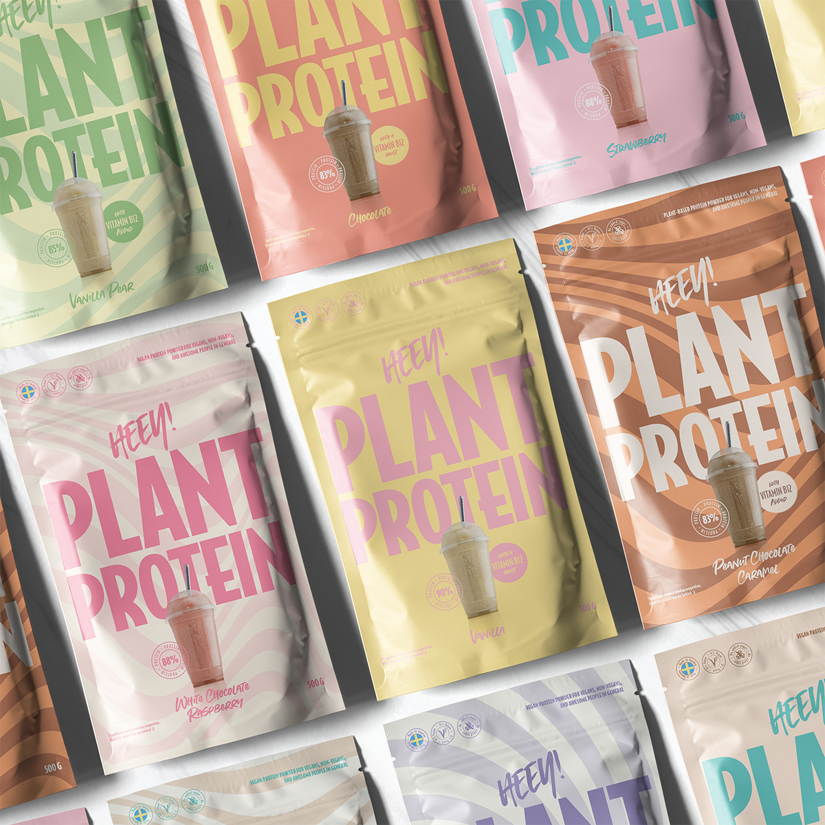 Several colorful plant protein powder bags are arranged in a grid. Packaging is matte with different solid or gradient colors per bag, mostly pinks and yellows. Bags are rectangular with rounded tops and resealable closures. Large sans-serif text reads “PLANT PROTEIN.” Smaller texts indicate flavors such as vanilla rose and strawberry. Ingredient lists and simple line-drawn shake illustrations appear on each bag. Background is plain white; no people or hands visible in this studio product shot.