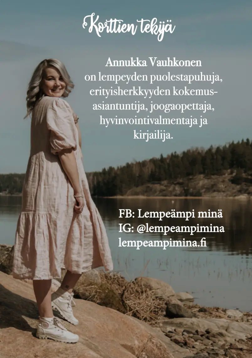Hidasta Elämää Lempeästi itseni puolella -kortit: vaaleaan mekkoon pukeutunut nainen seisoo hymyillen laiturilla järven rannalla; suuri, luettava teksti esittelee Annukka Vauhkosen hyvinvointivalmentajana, kirjailijana ja muuna; sosiaalisen median yhteystiedot näkyvät pienemmällä fontilla; ympäristö on ulkona veden äärellä luonnonvalossa; pakkausta tai tuoteastioita ei näy kuvassa; hyvinvointi ja tasapaino ovat hänen lähestymistapansa ytimessä.