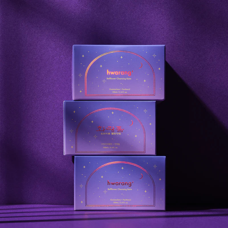 Hwarang Bellflower Cleansing Balm; kolme violettia Hwarang Safflower Cleansing Balm -rasiaa pinottuna mattapintaista violettia taustaa vasten; suorakaiteen muotoisissa rasioissa kultaisia tähtikuvioita, kuunsirppi, saflorikukkien kuvituksia ja koreankielistä tekstiä lihavoidulla valkoisella ja kultaisella fontilla; rasioiden varjot ulottuvat vasemmalle; ei ihmisiä tai käsiä kuvassa.