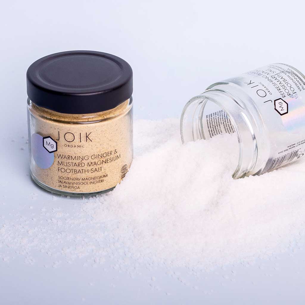 JOIK Organic Warming Magnesium Jalkakylpysuola; kaksi lasipurkkia, merkitty Warming Ginger & Mustard Magnesium Footbath-Salt, toinen pystyssä ja toinen kaatuneena, näyttävät rauhoittavaa magnesiumkylpysuolaa valuneena valkoiselle pinnalle; kirkkaat sylinterinmuotoiset lasiastiat mustin kierrekorkein; sisältö on karkeaa murretun valkoista kidettä; etiketeissä pelkistettyä mustaa tekstiä mattapintaisella valkoisella paperilla; tausta puhdas ja yksivärinen valkoinen; ei ihmisiä tai käsiä näkyvissä.