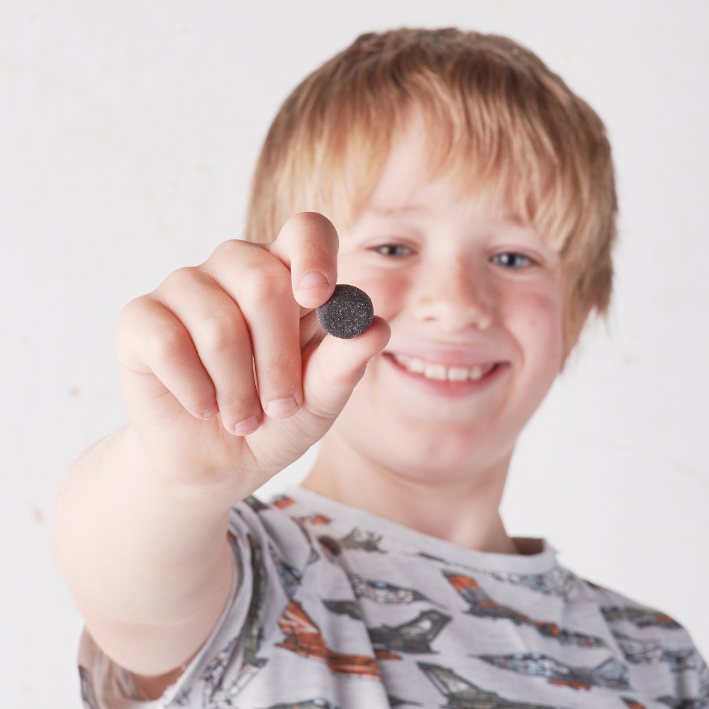 Kiki Health Body Biotics Gummies Maitohappobakteeri; hymyilevä, vaaleahiuksinen lapsi pitelee pientä pyöreää mustaa esinettä kohti kameraa, yllään paita, jossa värikkäitä lentokoneita, valkoista puhdasta taustaa vasten; esine on täysin musta ja vaikuttaa sileältä ja mattapintaiselta ilman näkyvää pakkausta tai etikettiä; kohtauksessa ei rekvisiittaa tai muita elementtejä, kuva rajattu tiiviisti yläkehoon ja tuotteeseen.