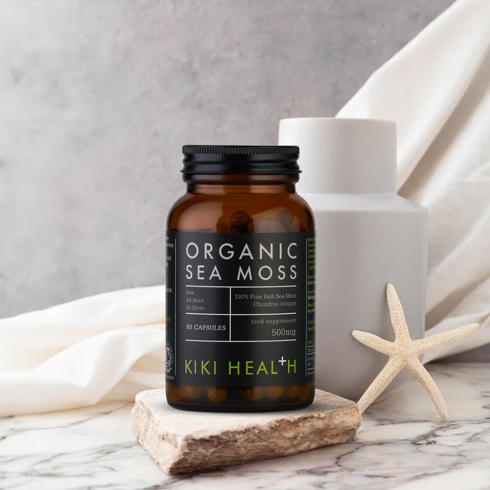 Kiki Health Irish Sea Moss; purkki KIKI Health Organic Sea Moss -kapseleita seisoo valkoisella marmoripinnalla; purkki on tummanruskeaa lasia mustalla mattapintaisella kierrekorkilla, etiketissä lihava groteskiteksti ”KIKI Health Organic Sea Moss Capsules”; lähellä sileitä harmaita ja beigejä kiviä, vaalea meritähti ja minimalistisia sisustuselementtejä taustalla; ei ihmisiä tai käsiä näkyvissä.