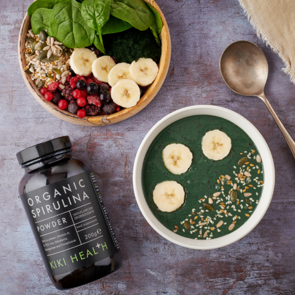 Kiki Health Luomu Spirulina-jauhe; kulho vihreää smoothieta, jota on rikastettu luomuspirulinajauheella, päällä banaaniviipaleita ja siemeniä, lähellä kulhoa hedelmiä, siemeniä ja pinaattia; pullo, merkitty Organic Spirulina Powder, ja lusikka harmaalla pinnalla lähellä; molemmat kulhot ovat pyöreitä, pullo sylinterinmuotoista muovia mattaetikettein groteskitekstillä; ei ihmisiä tai käsiä näkyvissä; kuva otettu ylhäältä.