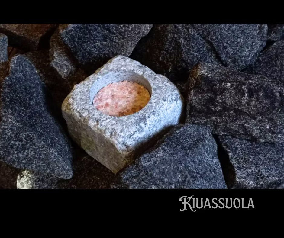 Kiuassuola ILMARI Aloituspakkaus; vaaleanpunaiskeskinen graniittikuutio tummien, epäsäännöllisten kivien joukossa; graniitti on mattapintainen ja kuutionmuotoinen; pakkausta ei näy kuvassa; tausta koostuu löyhästi levitetyistä tummista, epätasaisista kivistä; "Kiuassuola" kirjoitettu selkeällä groteskilla kuvan oikeassa alakulmassa; ei ihmisiä tai käsiä kuvassa.