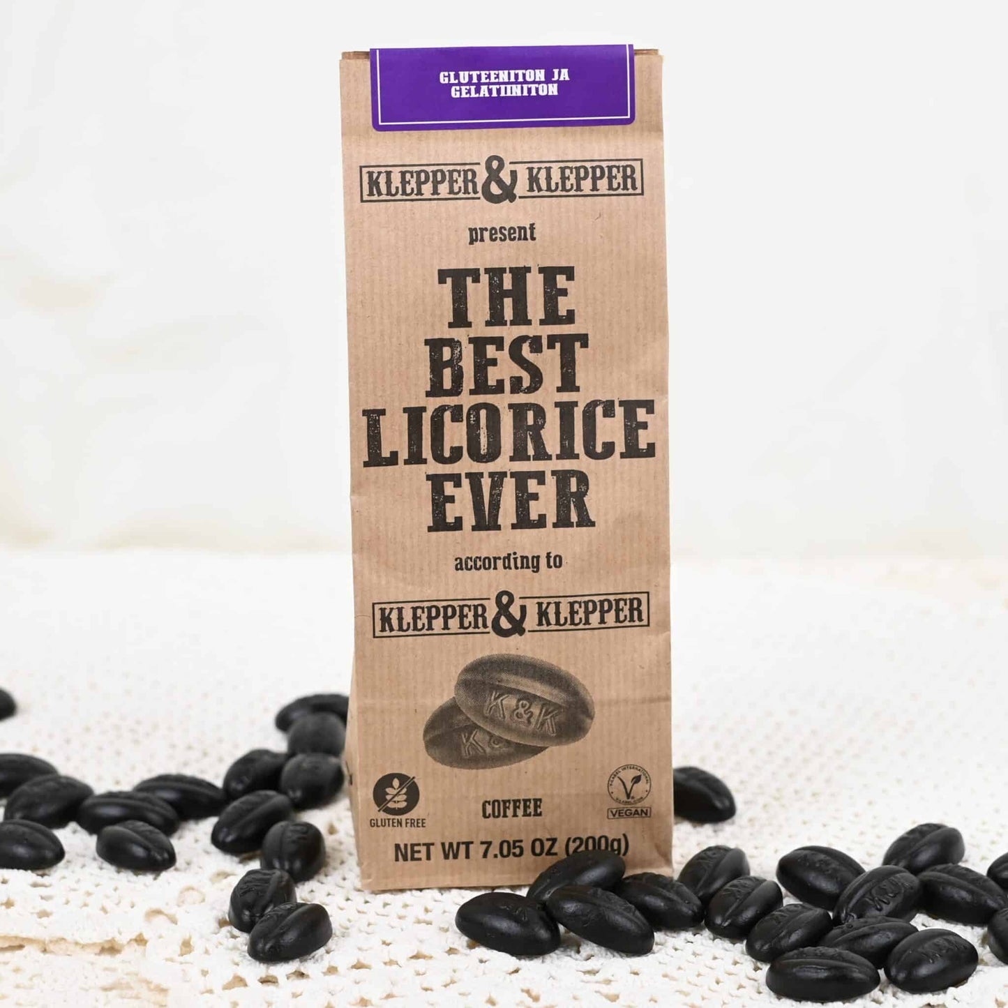 Klepper & Klepper Gluteeniton Lakritsi Coffee; ruskea paperipussi tekstillä ”The Best Licorice Ever by Klepper & Klepper” valkoisella alustalla; pussi mattapintainen ja pystyssä, painettua mustaa tekstiä sekaisin antiikva- ja groteskifonteilla; pussin ympärillä kiiltäviä mustia lakritsipaloja kahvipavun muodossa; ei ihmisiä tai käsiä näkyvissä; kuva otettu hieman viistosti ylhäältä.