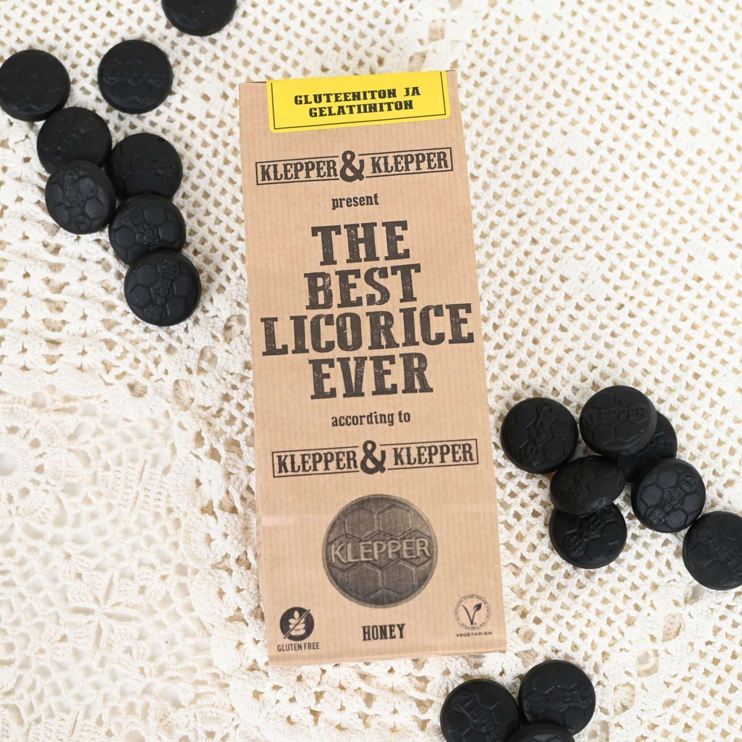Klepper & Klepper Gluteeniton Lakritsi Honey; suorakaiteen muotoinen pakkaus tekstillä The Best Licorice Ever on aseteltu valkoiselle pitsiliinalle, pyöreitä mustia lakritsipaloja ympärillä; pakkauksessa lihavoitua groteskitekstiä kertoen gluteenittomasta, liivatteettomasta ja hunajan makuisesta lakritsista; ei ihmisiä tai käsiä näkyvissä; kuva otettu yläviistosta.