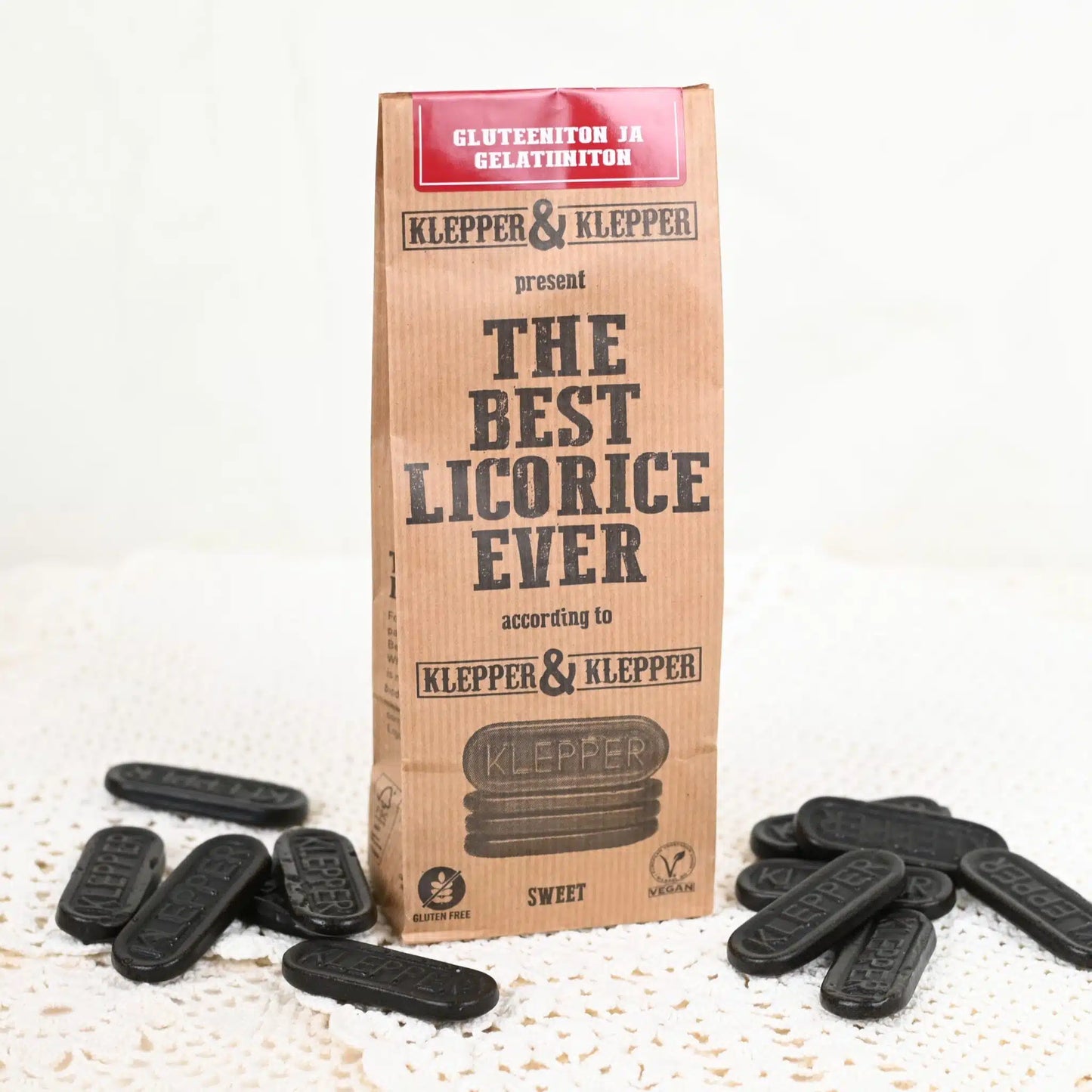 A rectangular package labeled The Best Licorice Ever stands upright on a white lace cloth. Several pieces of glossy black licorice are scattered around it. Packaging is mainly matte, with bold sans-serif text and smaller details noting gluten-free and vegan. No people or hands present; image is a product shot. A package labeled The Best Licorice Ever stands on a lace cloth, surrounded by pieces of black licorice. The gluten-free, vegan packaging highlights its irresistibly sweet flavor.