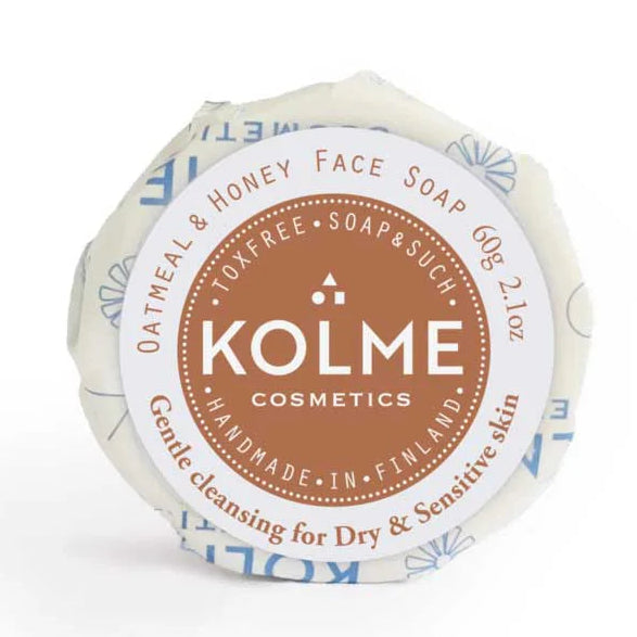 Kolme Cosmetics Kasvosaippua Kaura & Hunaja: pyöreä saippuapalanen käärittynä läpinäkymättömään valkoiseen paperiin; etiketti on ruskea ja valkoinen siistillä groteskikirjoituksella; etiketissä mainitaan käsintehty Suomessa ja kauran sisältö kuivalle ja herkälle iholle; ei käsiä tai ihmisiä näkyvissä; kuva on lähikuva neutraalia taustaa vasten.