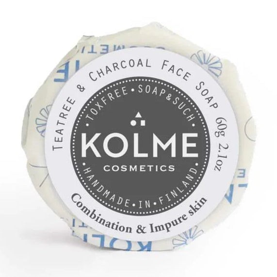 Kolme Cosmetics Kasvosaippua Teepuuöljy & Aktiivihiili: pyöreä saippuapalanen käärittynä valkoiseen paperiin sinisin kuvioin; ympyränmuotoisessa etiketissä groteskikirjasin ja teksti: "KOLME COSMETICS Teatree & Charcoal Face Soap"; käsintehty Suomessa yhdistelmä- ja epäpuhtaalle iholle, ainutlaatuinen kaava hyödyntää aktiivihiilen puhdistavia ominaisuuksia; tuote kuvattu yksinään neutraalia taustaa vasten, ei ihmisiä tai käsiä näkyvissä.