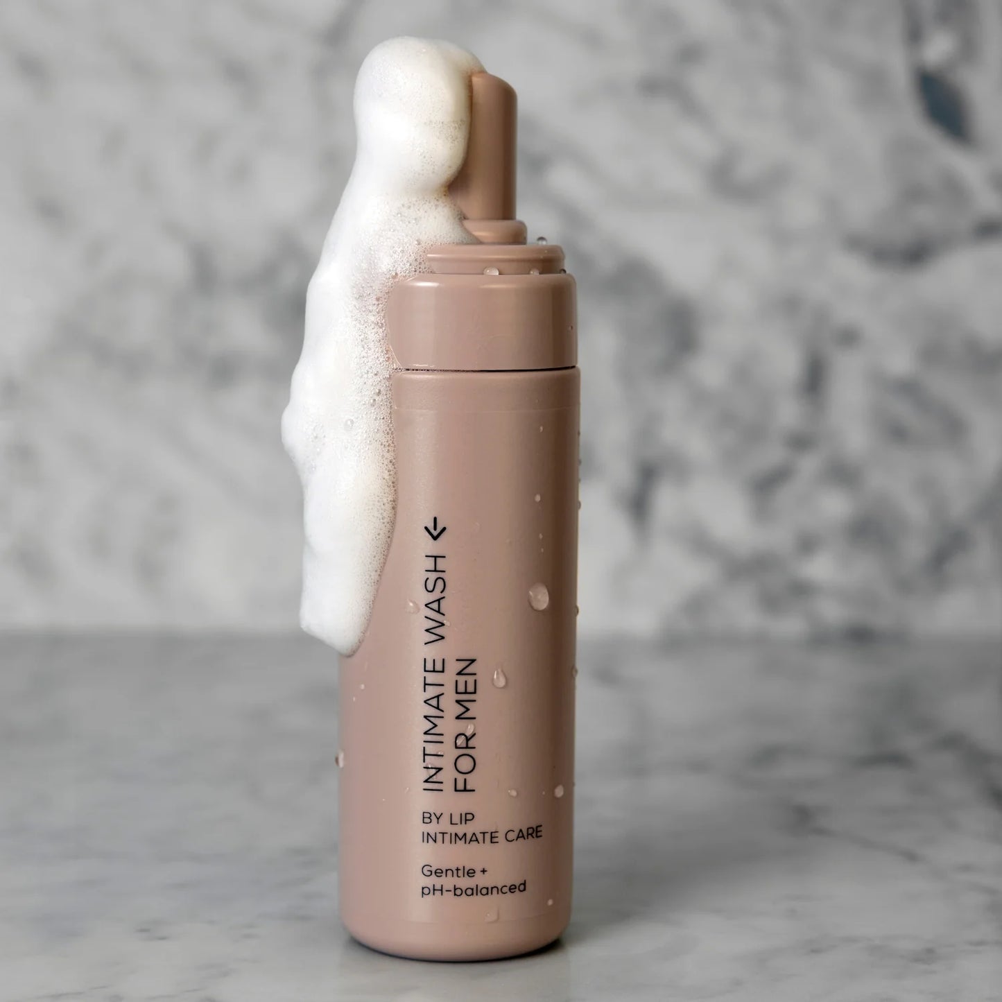 LIP Intiimipesuaine Miehille; beige pullo, jossa lukee "Intimate Wash for Men", seisoo marmoripinnalla, valkoista vaahtoa valuu mattapintaisesta annostelijasta yli; etiketin teksti on yksinkertaista, mustaa ja groteskia; suorakulmaisessa pullossa on pyöristetyt reunat eikä näkyviä kuvioita; tausta on harmaa ja marmoroitu läpi kuvan, kuvassa ei ihmisiä tai käsiä.