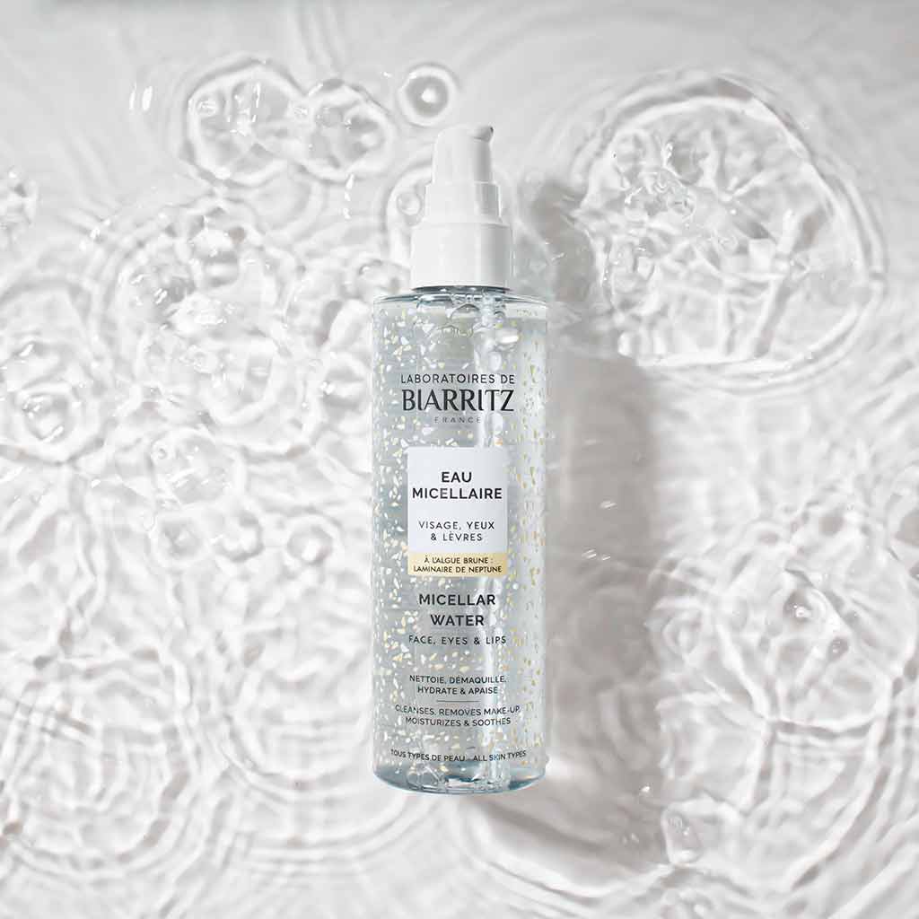 Laboratoires de Biarritz Cleansing Care Misellivesi; kirkas muovipullo tekstillä "Laboratoires de Biarritz Micellar Water" pystyssä lievästi väreilevän veden pinnalla; lieriön muotoinen pullo valkoisella litteällä korkilla ja kiiltäväpintaisena; etiketissä yksinkertainen musta groteskiteksti sekä sinisiä ja turkoosinvärisiä korostuksia; ei ihmisiä tai käsiä; studiovalaistus luo heijastuksia veteen ja pulloon.