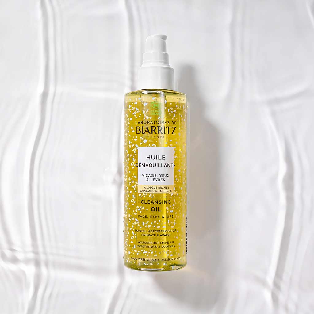 Laboratoires de Biarritz Cleansing Care Puhdistusöljy; pullo pumppuannostelijalla asetettuna tekstuuriselle valkoiselle pinnalle; kirkas lieriön muotoinen muovipullo kiiltäväpintaisena, sisältää keltaista nestettä valkoisin kuplin tai helmin; etusivun etiketissä tuotenimi tummin kirjaimin; ei ihmisiä tai käsiä kuvassa.