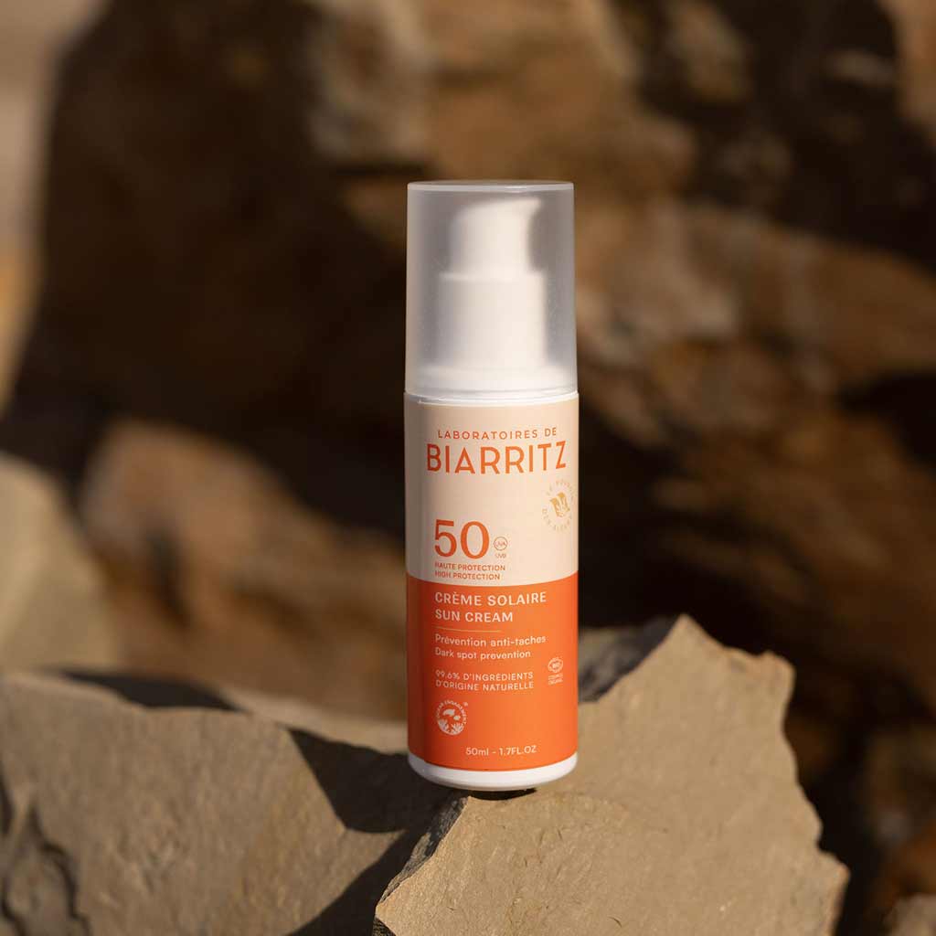 Laboratoires de Biarritz Face Sun Cream Aurinkovoide Kasvoille SPF50; pullo Laboratoires de Biarritz SPF 50 -kasvojen aurinkovoidetta lepää vaaleanruskealla kivellä, mineraalirikkaat ainesosat korostettuna epätarkkaa kivistä taustaa vasten lämpimässä auringonvalossa; pullo valkoista muovia oranssilla korkilla ja mattapinnalla; sylinterin muotoinen pullo tekstillä brändi ja SPF 50 mustana ja oranssina; ei ihmisiä tai käsiä näkyvissä.