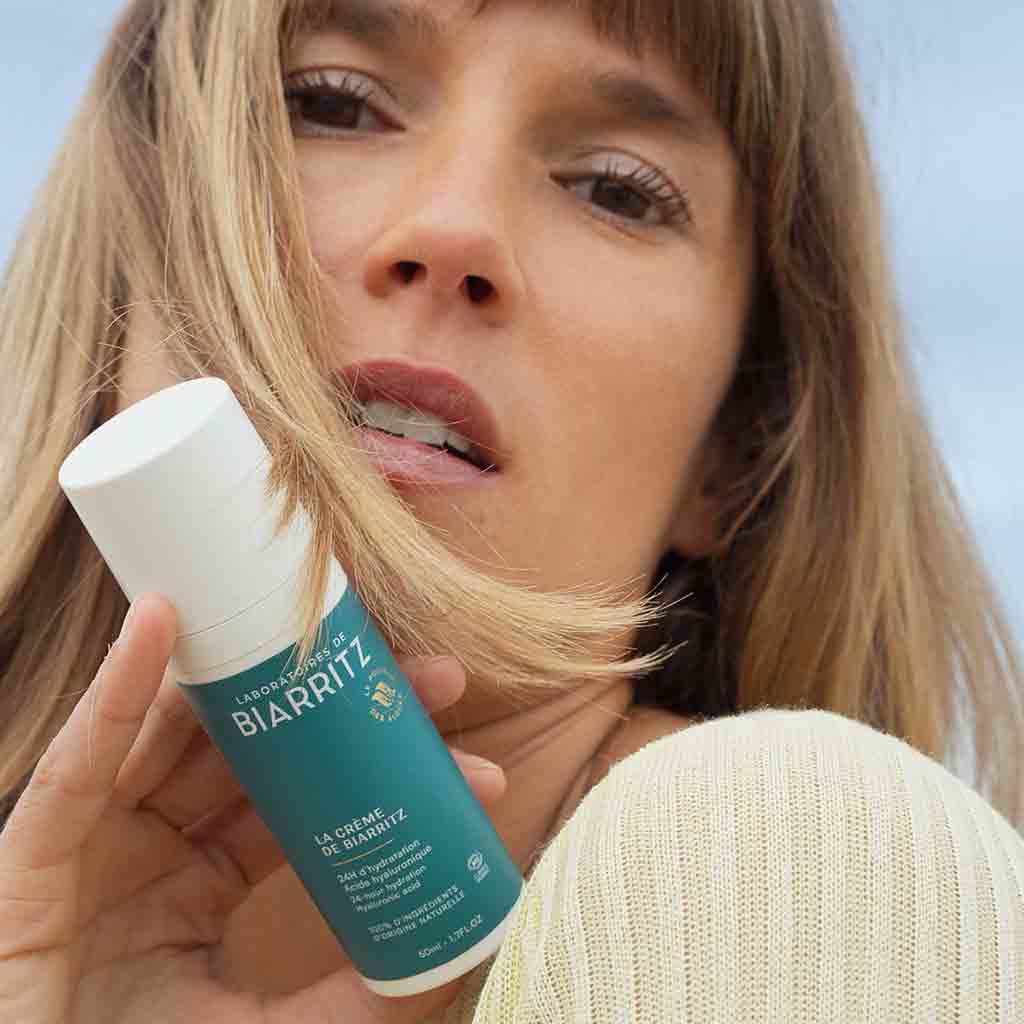 Laboratoires de Biarritz La Crème de Biarritz Face Cream: vaaleahiuksinen nainen pitelee kasvojensa lähellä sylinterimäistä valkoista pulloa, jossa on pumppuannostelija ja mattapintainen etiketti; etiketissä tumma groteskiteksti "Laboratoires Biarritz" ja sinisiä korostuksia; nainen katsoo kameraan; tausta on kirkas, vaaleansininen taivas.