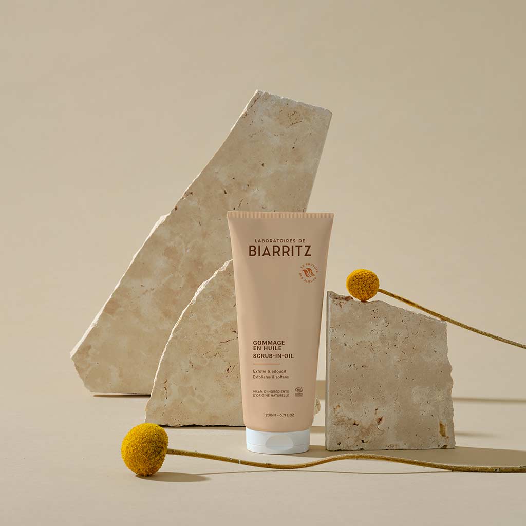 Laboratoires de Biarritz Oil-In Body Scrub Vartalokuorinta; tuubi Laboratoires de Biarritz -öljykuorintaa seisoo pystyssä kahden beigen kivilaatan välissä, seurassaan kaksi keltaista pallukkakukkaa neutraalilla taustalla; valkoinen tuubi kiiltopinnalla ja läppäkorkilla; mustaa ja vihreää tekstiä nykyaikaisella groteskikirjasimella painettu pystysuuntaisesti etikettiin, ”Laboratoires de Biarritz” selkeästi näkyvissä; ei ihmisiä tai käsiä.