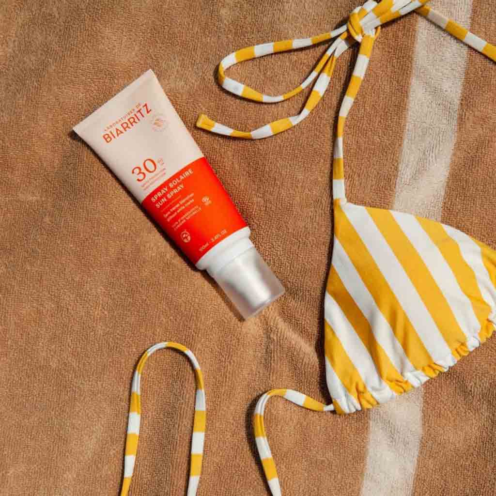 Laboratoires de Biarritz Sun Spray Aurinkosuojasuihke SPF30; tuubi SPF 30 -aurinkovoidetta valkoisella korkilla ja painetulla etiketillä aseteltuna vaalealle kangaspyyhkeelle; keltavalkoraidallinen bikiniyläosa sileästä kankaasta lepää sen vieressä; etiketissä ”SPF 30” lihavoidulla, selkeällä tekstillä; tuubin pinta puolikiiltävä; ei ihmisiä tai käsiä kuvassa.