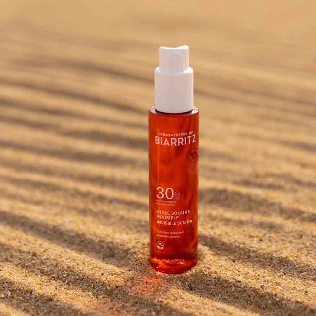 Laboratoires de Biarritz Suncare Invisible Oil SPF30; pullo Laboratoires de Biarritz SPF 30 -näkymätöntä aurinkoöljyä seisoo pystyssä hiekalla; pullo sylinterin muotoinen, mattavalkoinen ja läpikuulumaton; mustaa ja oranssia groteskitekstiä etiketissä lukee ”Laboratoires de Biarritz” ja ”SPF 30”; auringonvalo luo selkeitä varjoja tekstuuriselle vaalealle hiekalle; ei ihmisiä tai käsiä kuvassa.